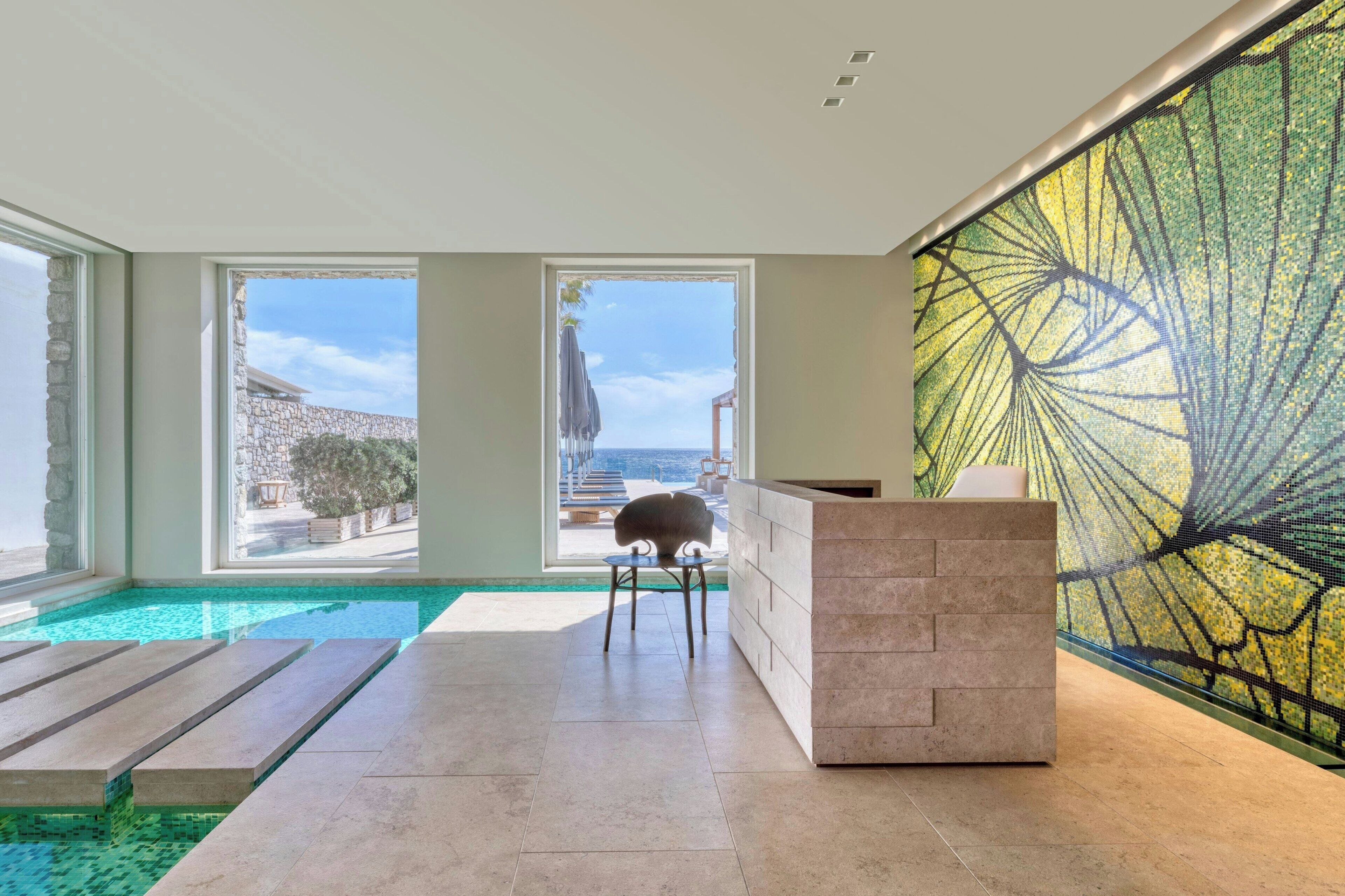 A modern indoor pool area with clear views of the sea and a stone wall outside.