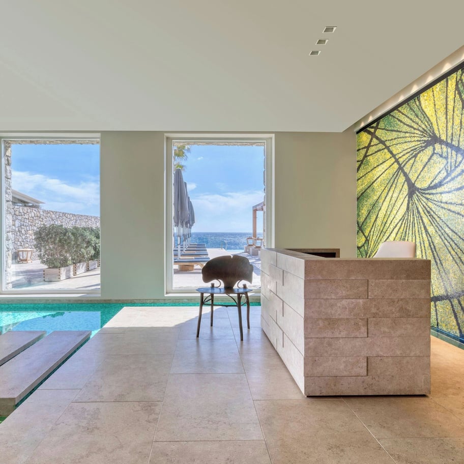 A modern indoor pool area with clear views of the sea and a stone wall outside.