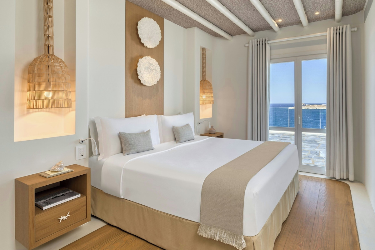 A modern bedroom with a large bed, bedside lamps, a nightstand, and a view of the sea through the window.