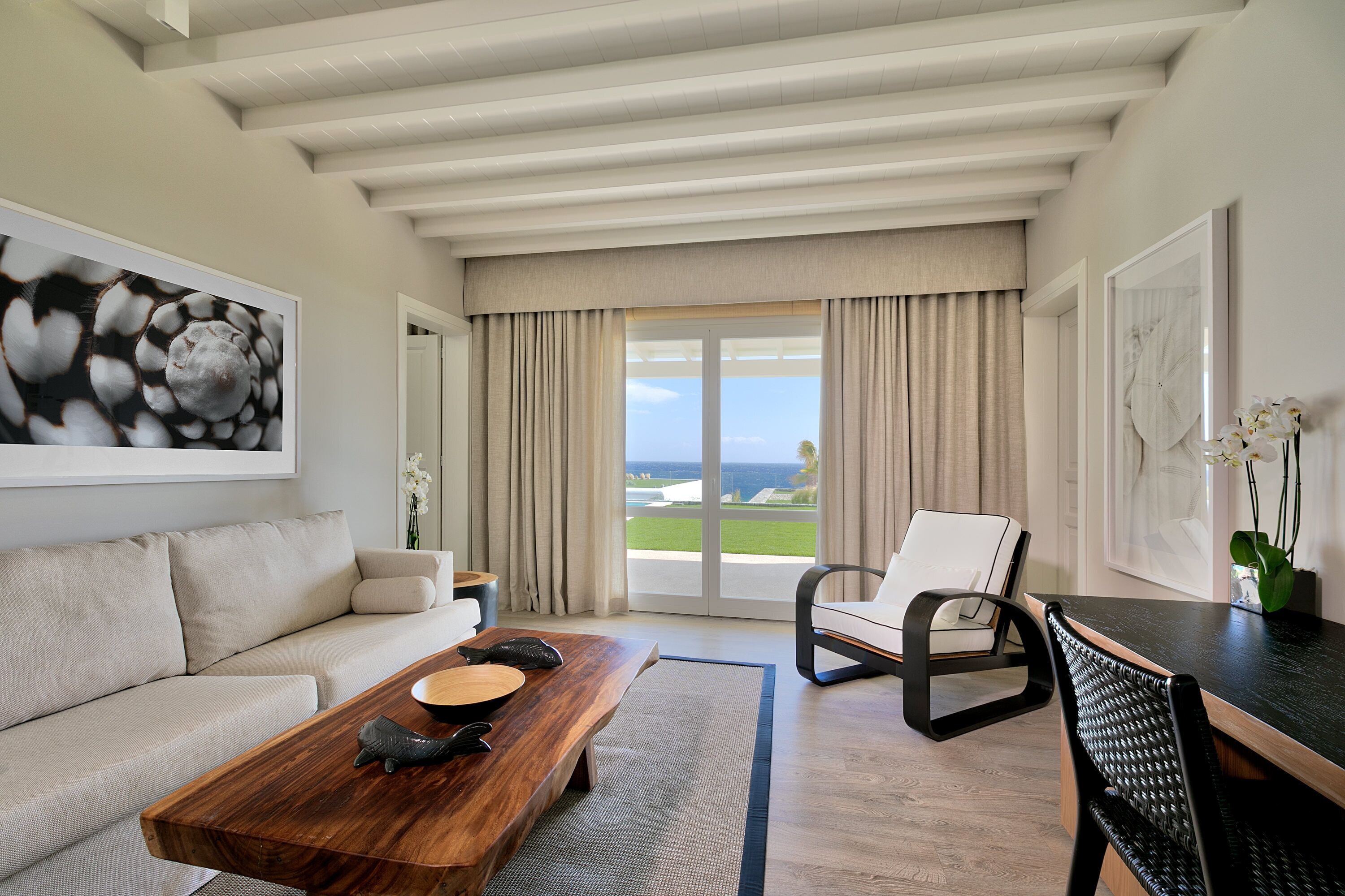 A modern living room with a wooden coffee table, a sofa, a chair, and a view of the ocean through large windows.
