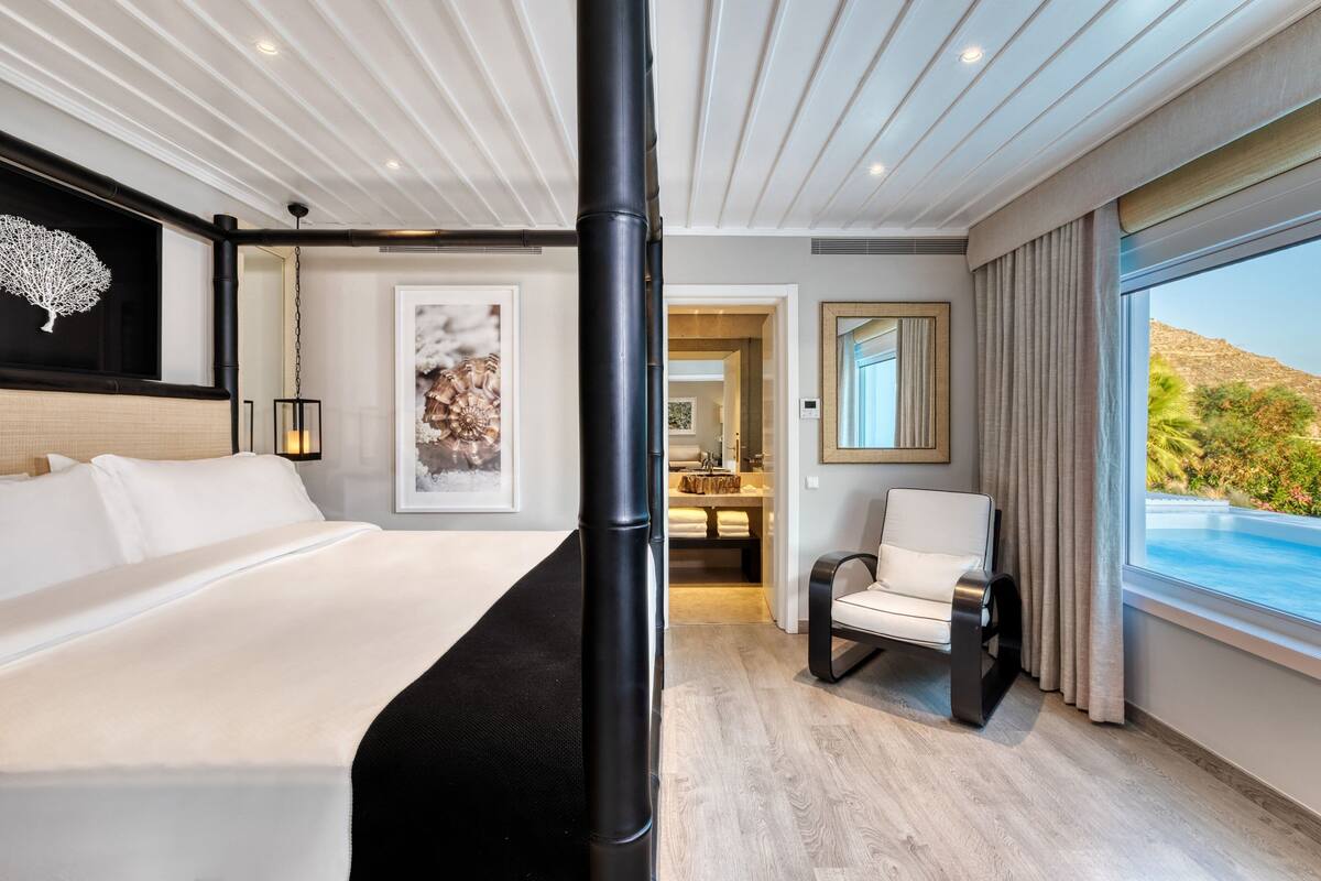 A modern hotel room with a large bed, a seating area, a view of a pool, and a dining area in the background.