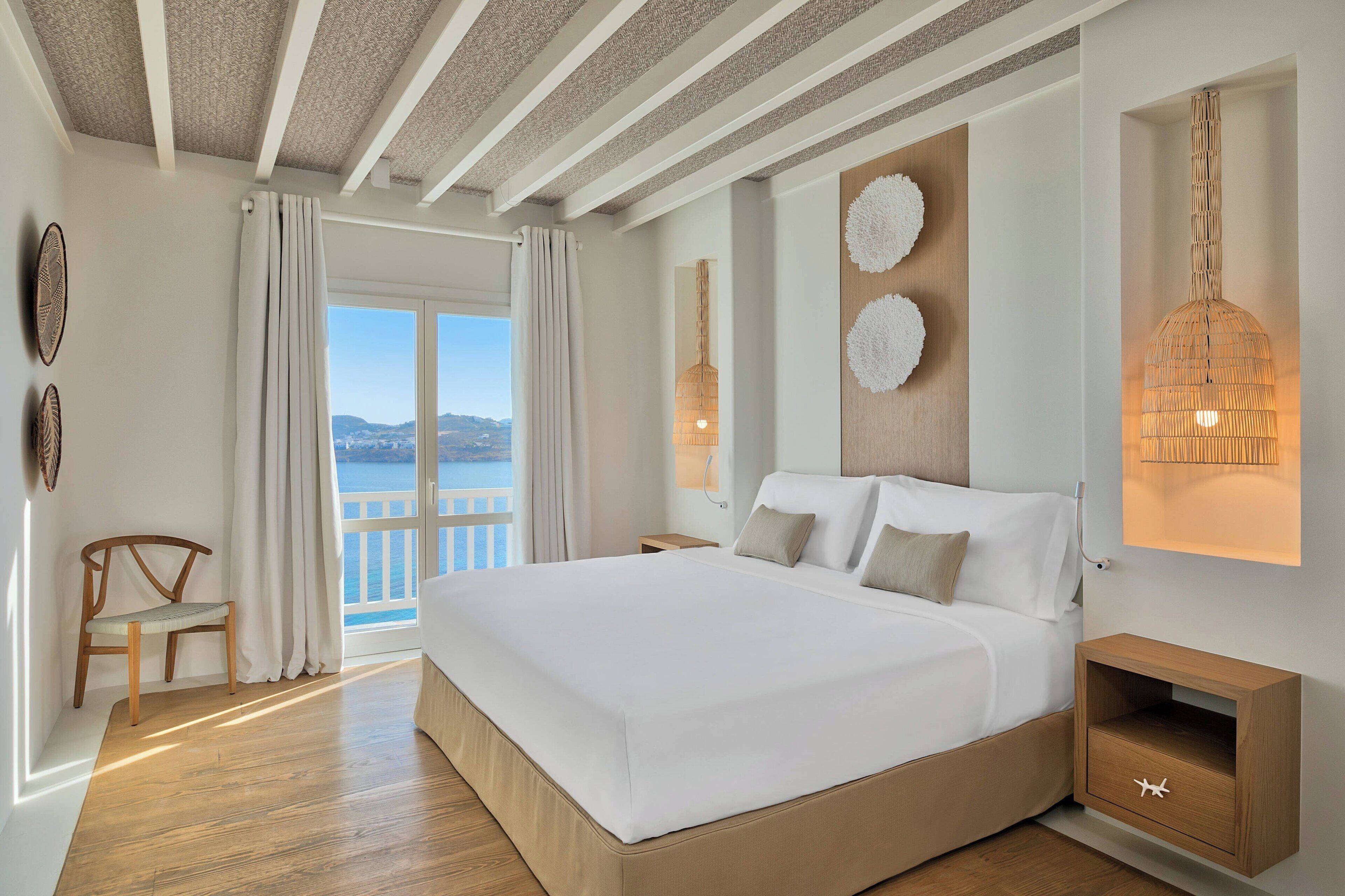 A modern bedroom with a large bed, a wooden nightstand, a chair, and a view of the sea.