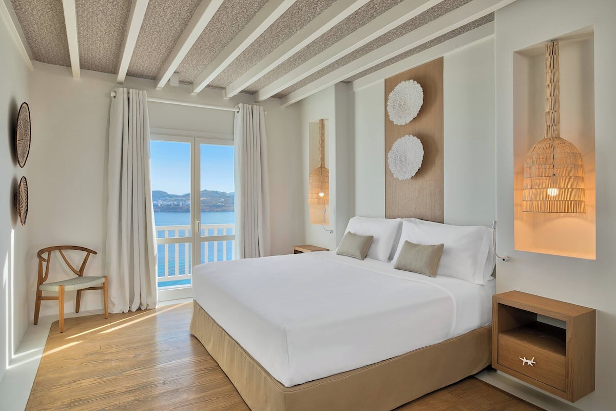 Quarto moderno com uma cama grande, mesa de cabeceira de madeira, uma cadeira e vista para o mar.