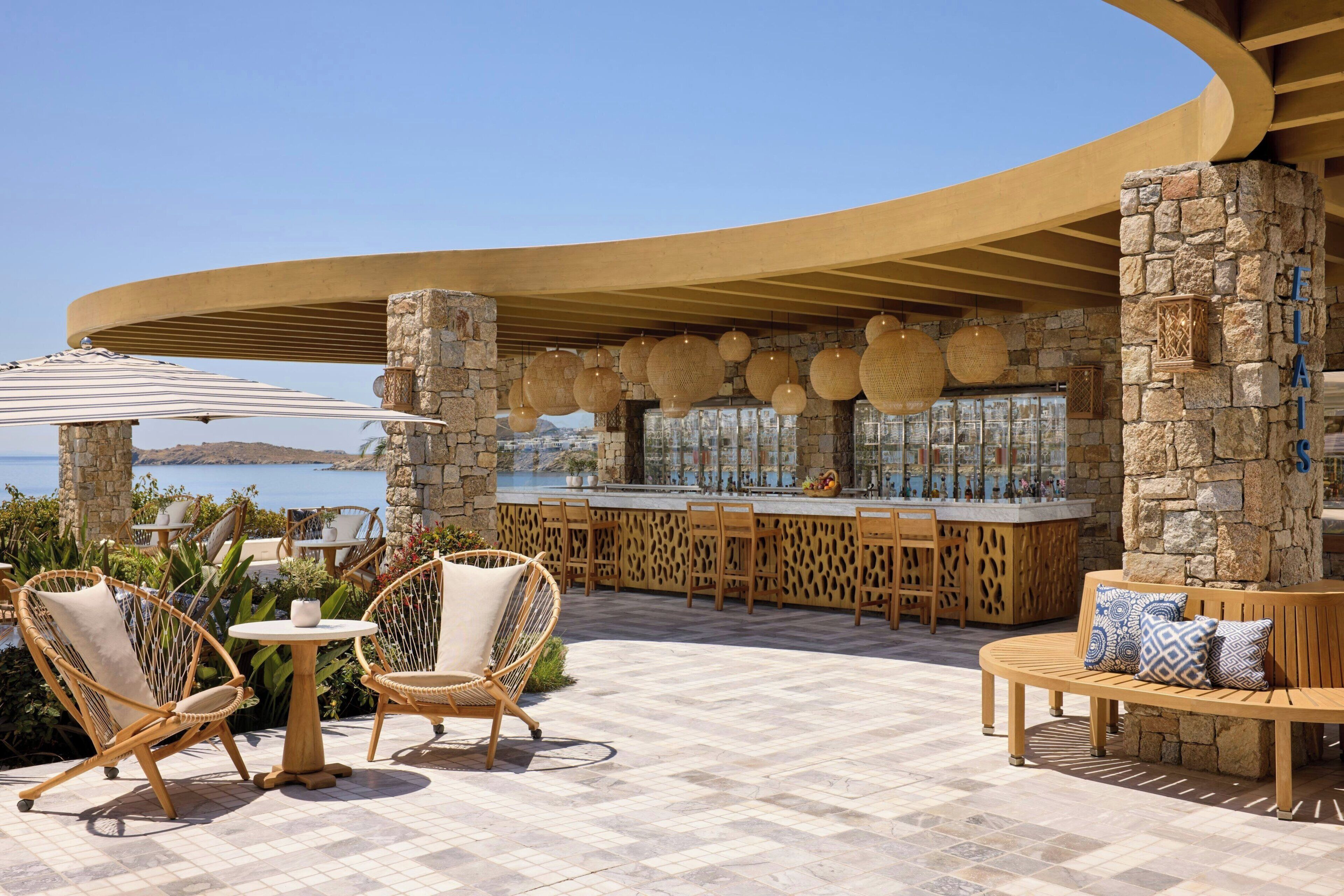A seaside bar area with wicker furniture, stone pillars, and a curved wooden ceiling.