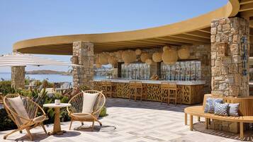 A seaside bar area with wicker furniture, stone pillars, and a curved wooden ceiling.