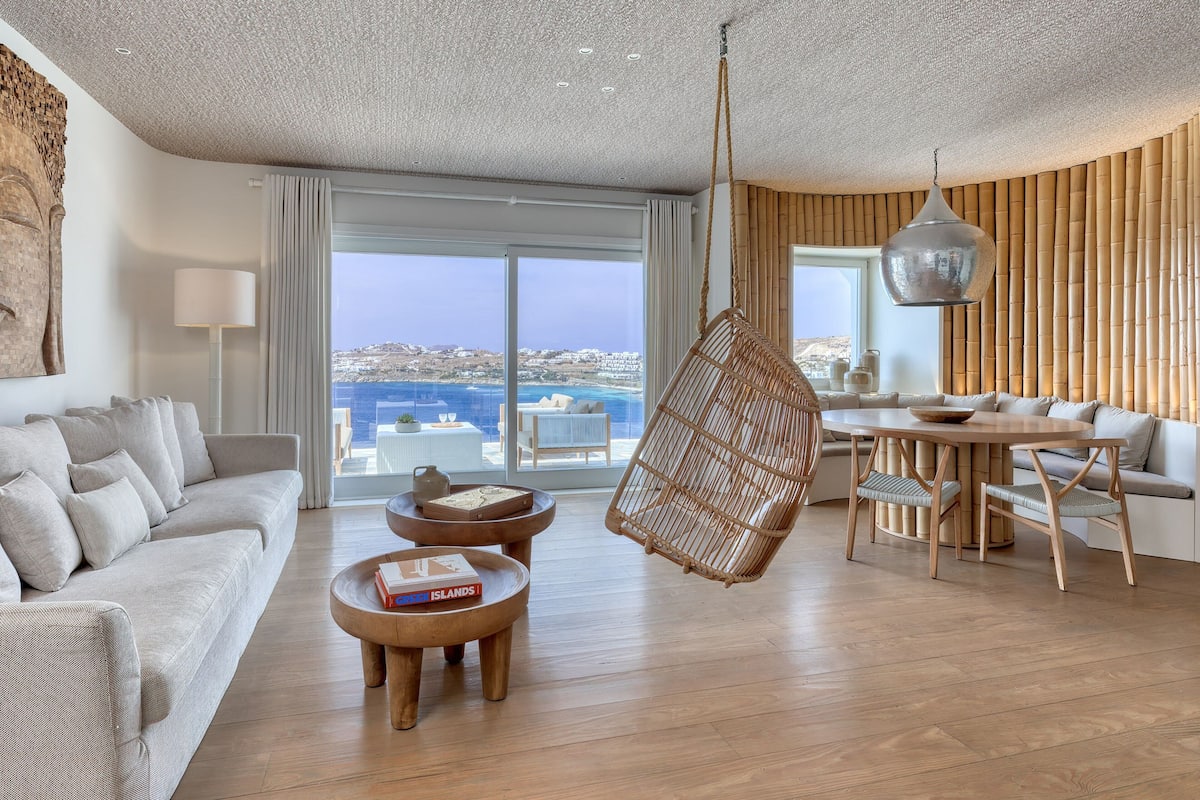Uma sala de estar moderna com sofá, duas mesas redondas de madeira, uma cadeira suspensa e um espaço de jantar com mesa e cadeiras.