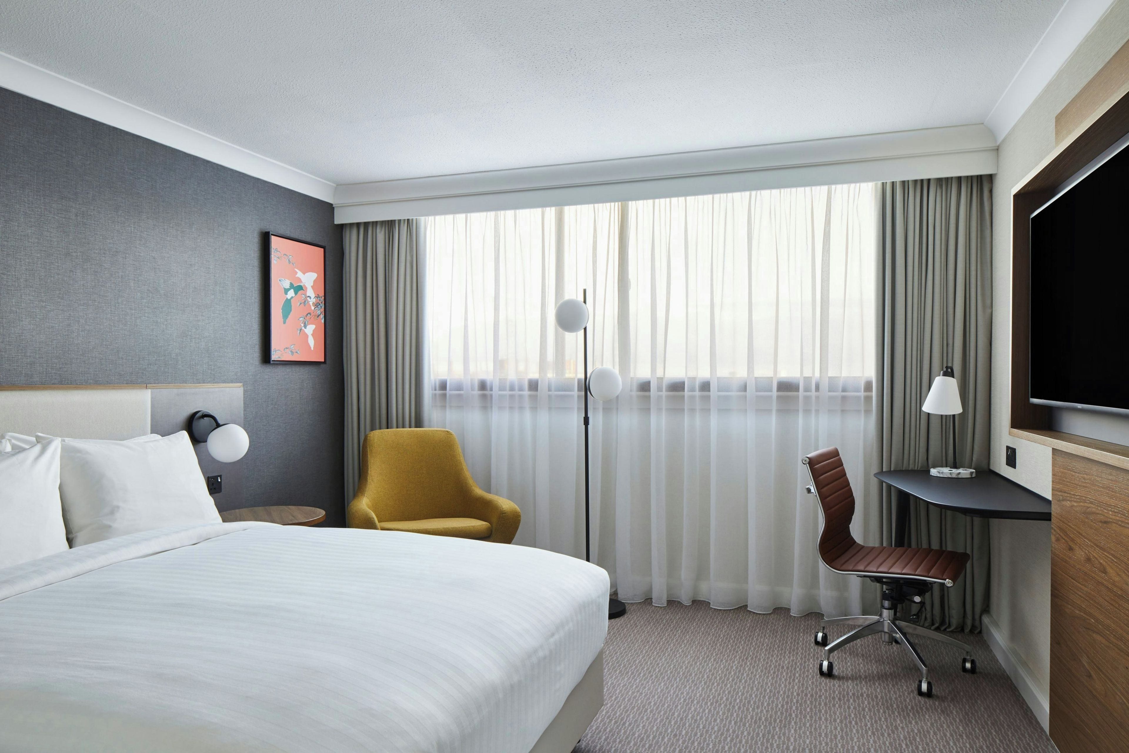 A hotel room with a bed, a desk, a chair, a TV, and a large window with curtains.