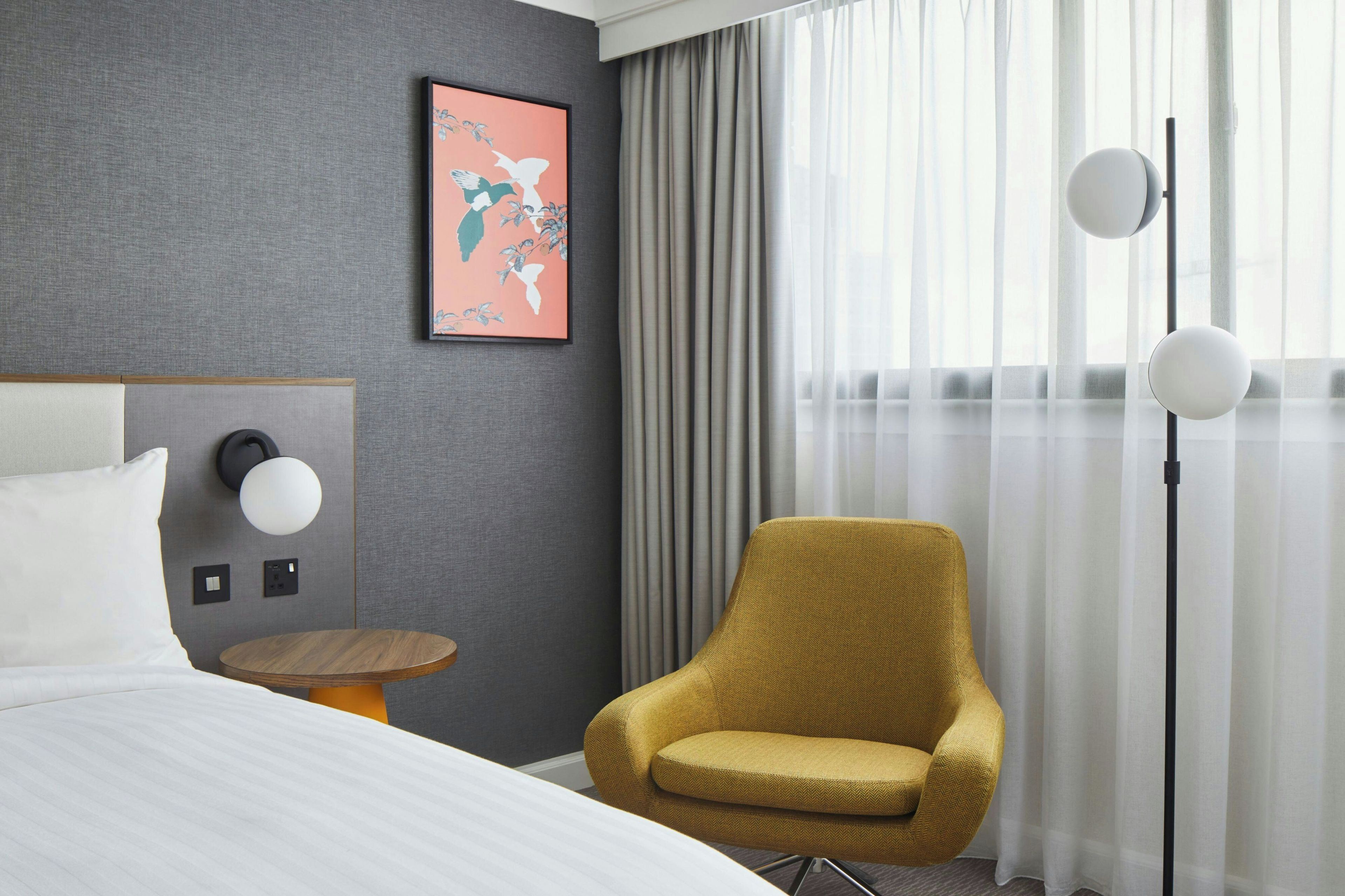 A hotel room with a bed, a round table, a chair, a floor lamp, and a window with curtains.