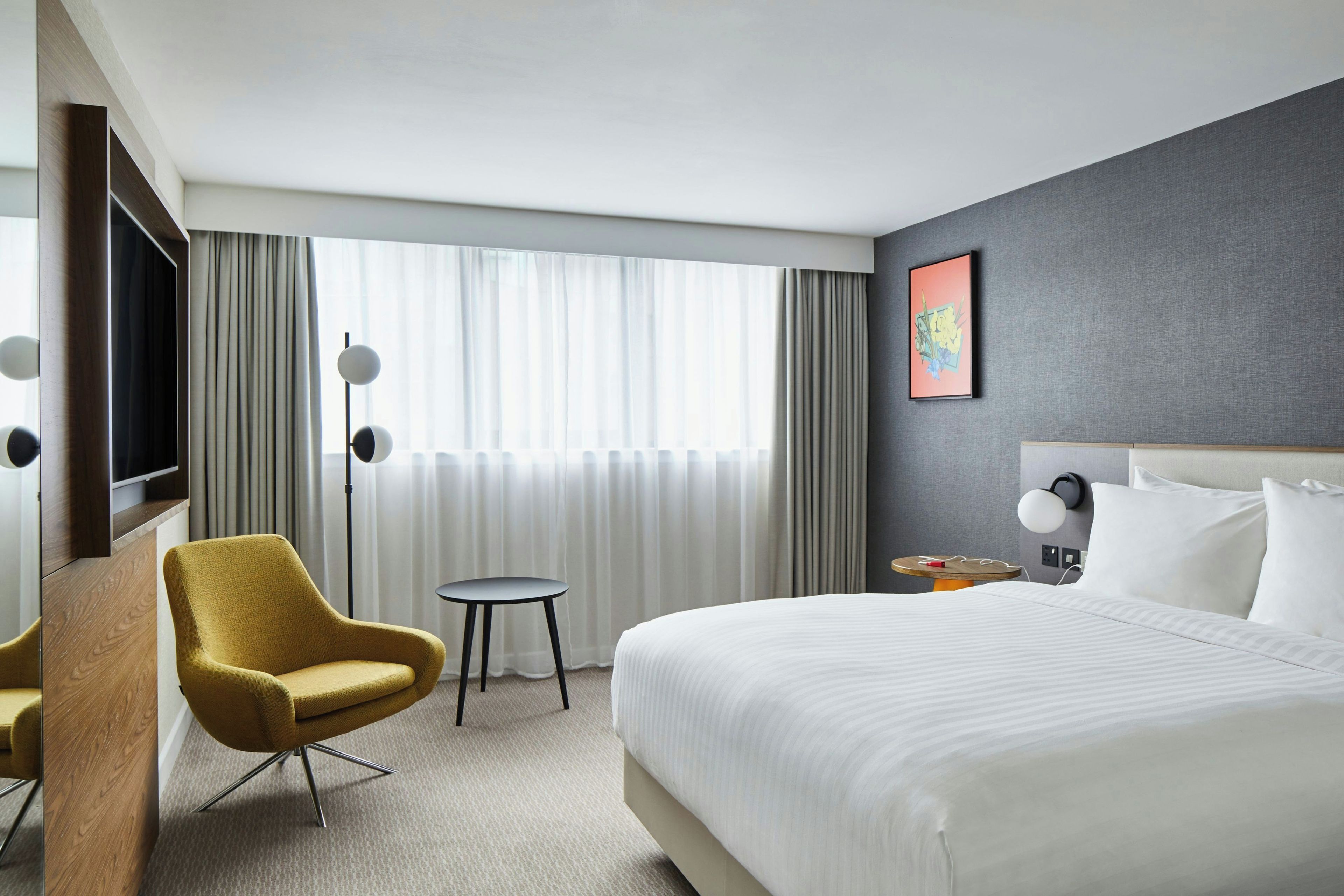 Una habitación de hotel con una cama, una silla, una mesita, una lámpara de pie y un cuadro en la pared.