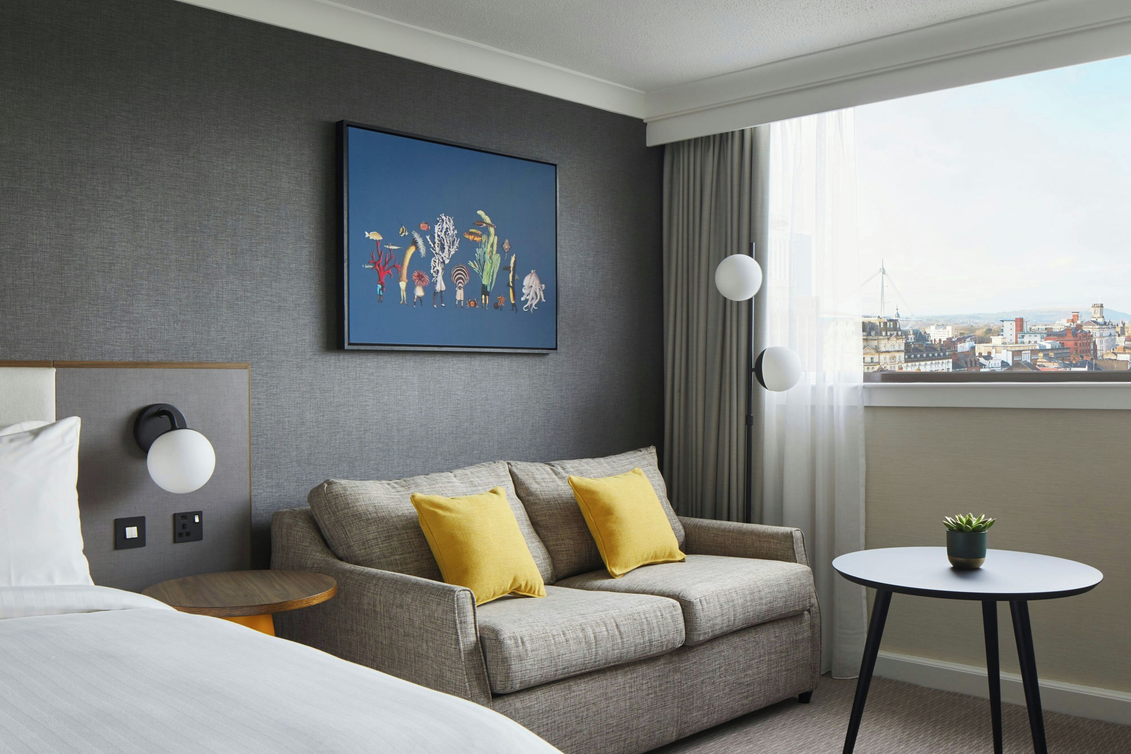 A hotel room with a grey sofa, two yellow cushions, a round table with a plant, a city view through the window, and a framed artwork on the wall.