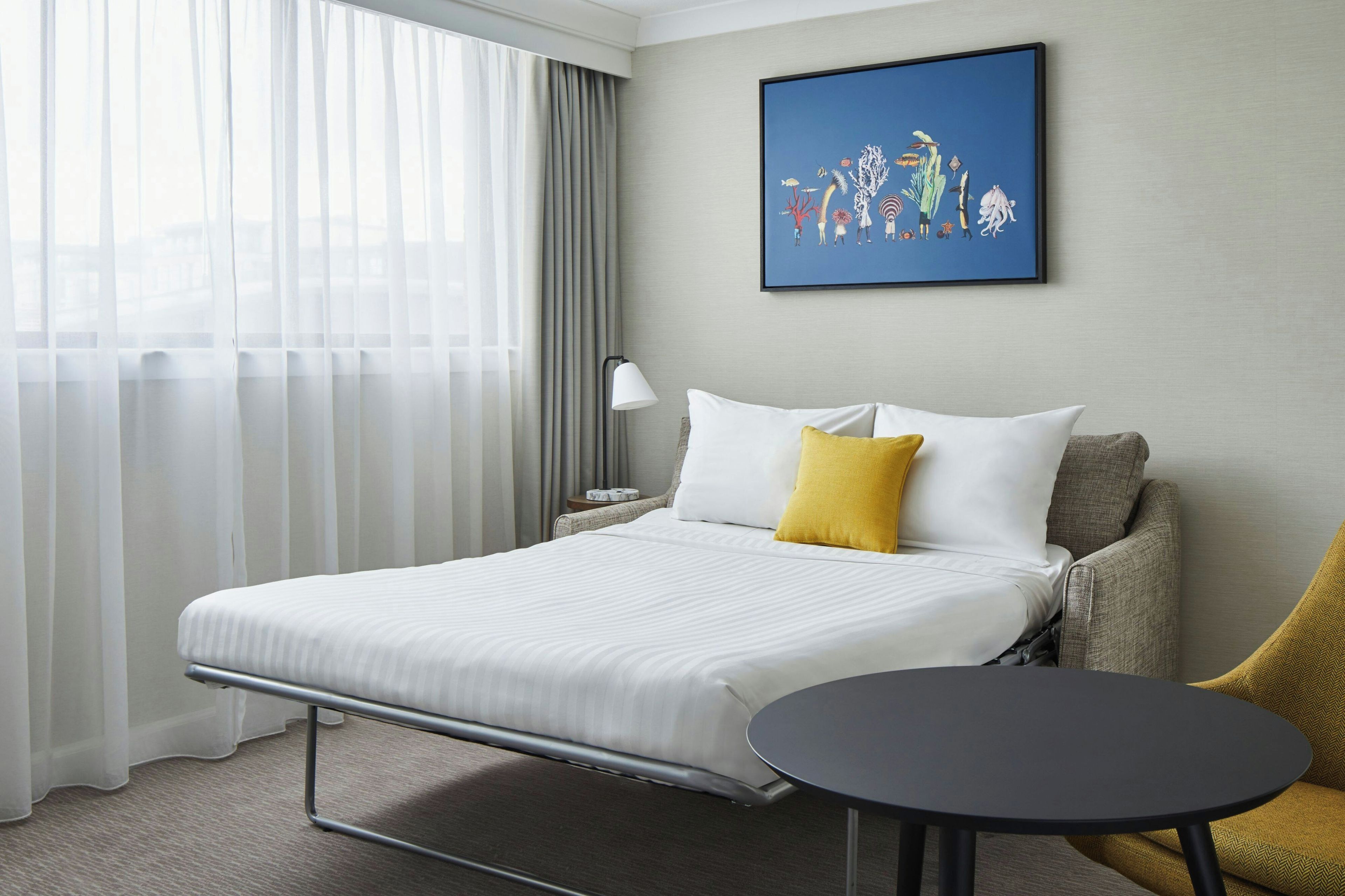 Une chambre d’hôtel avec un lit, une table de chevet, une chaise, une table ronde et un tableau accroché au mur.