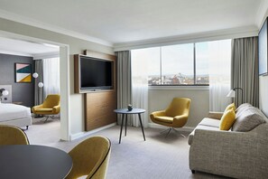 A hotel room with a bed, two armchairs, a sofa, a TV, a small table, and a view of the city.