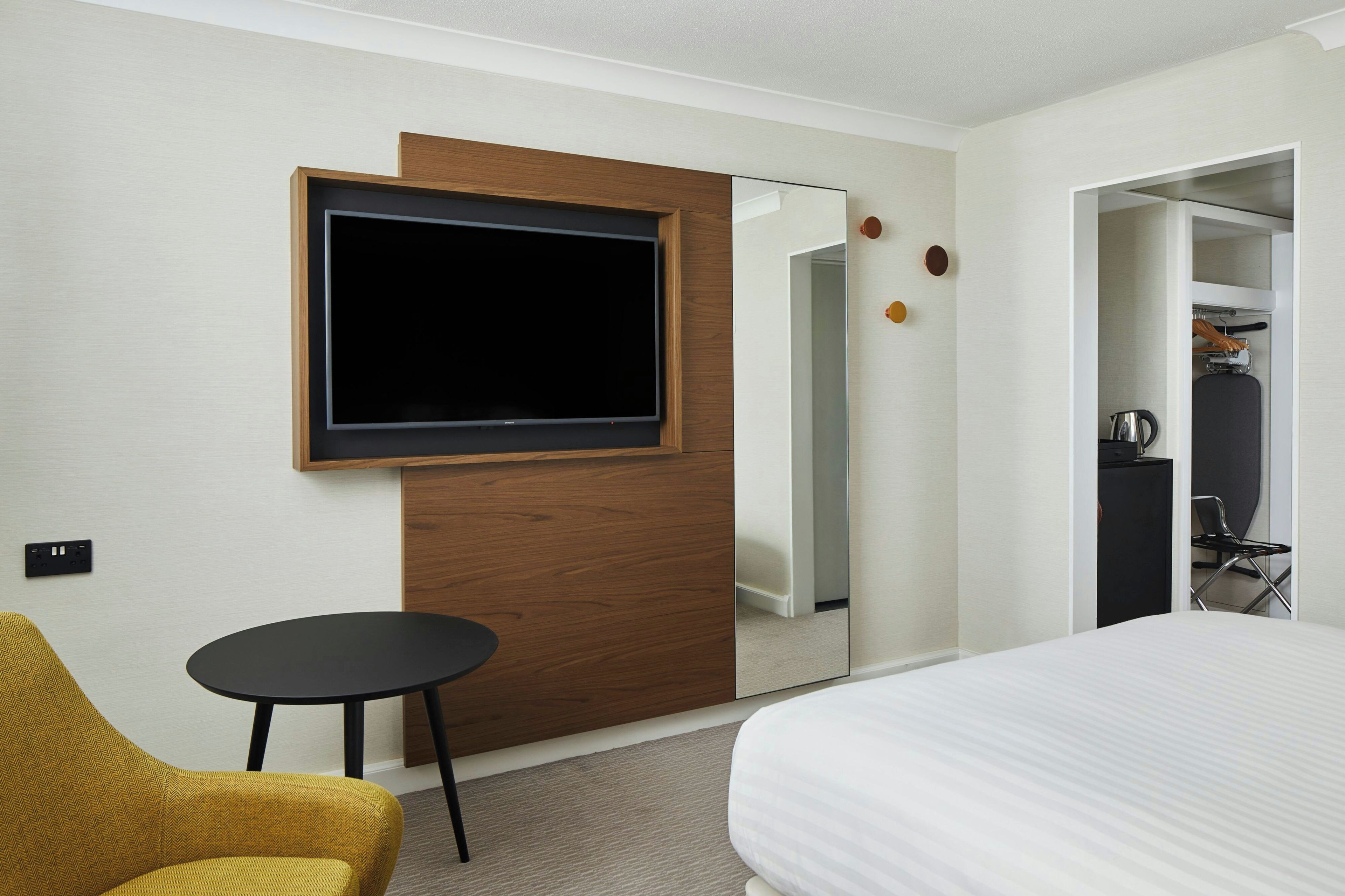 Une chambre d’hôtel avec un lit, un téléviseur fixé au mur, une petite table ronde et une chaise jaune.