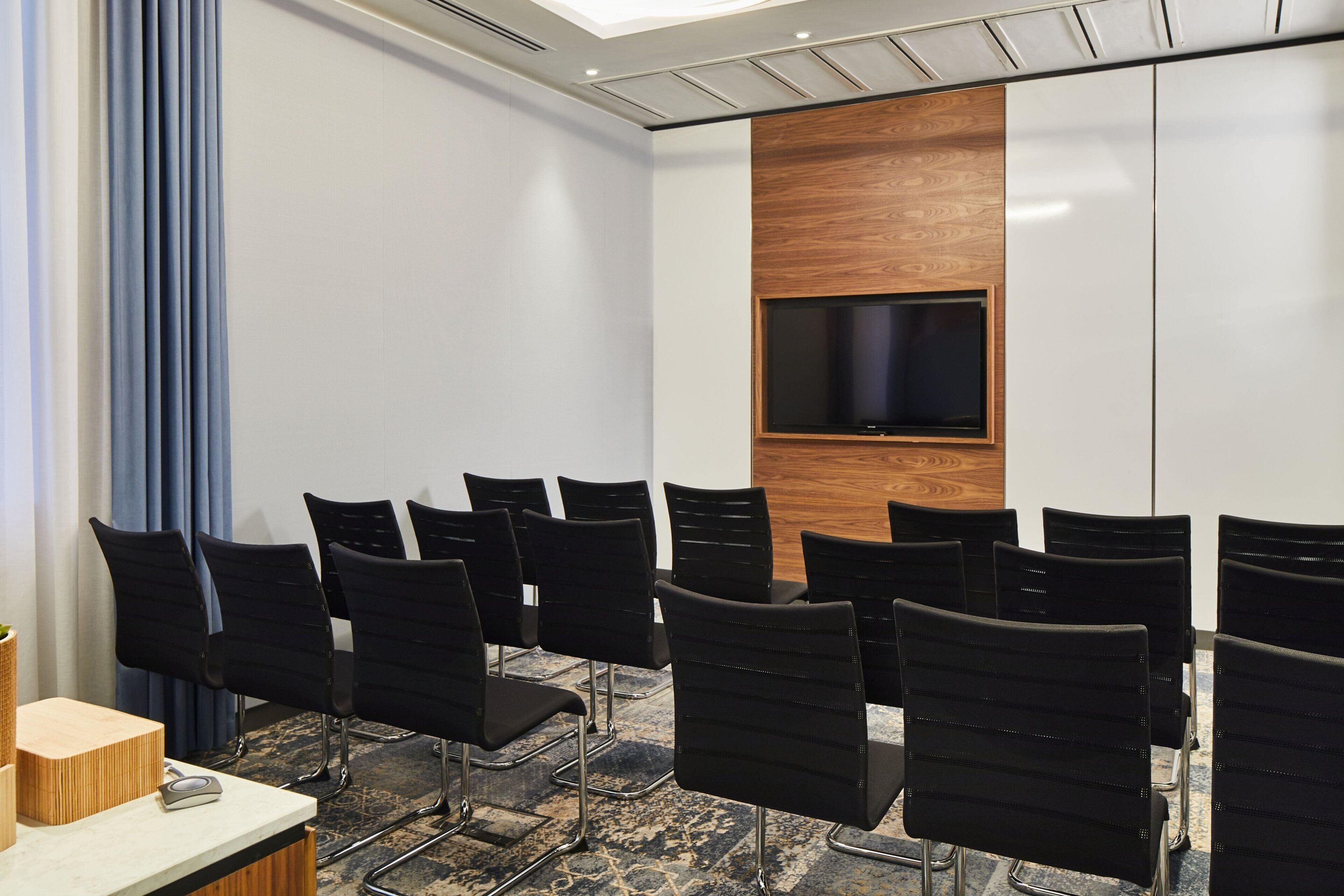 Uma sala de conferência com cadeiras pretas dispostas em fileiras, uma parede de destaque em madeira, uma TV de tela plana montada acima de uma mesa e um tapete estampado.