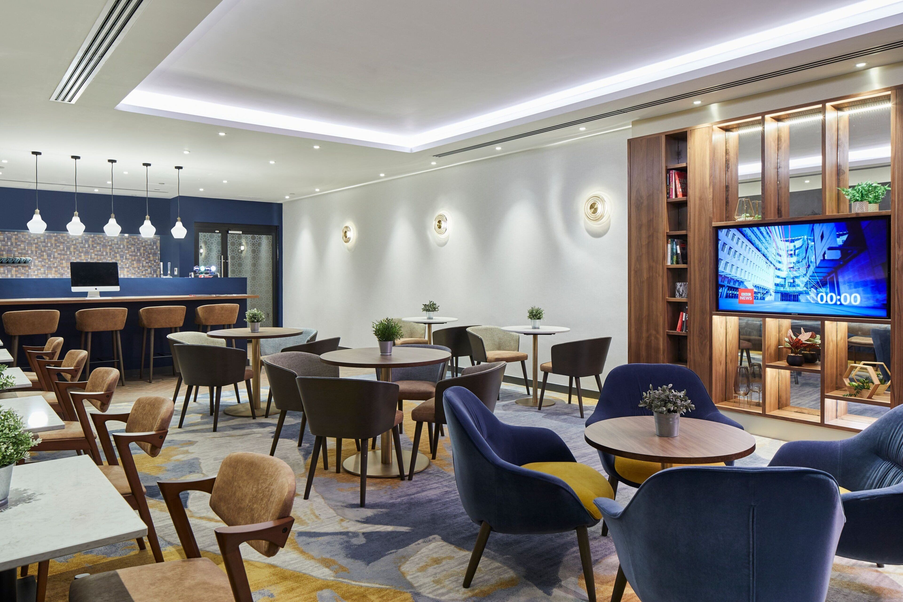 Un moderno vestíbulo de hotel con mesas redondas, asientos cómodos, un televisor y una zona de bar.
