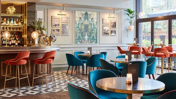 A bar area with a wooden counter, red bar stools, and a variety of bottles. There are blue chairs and tables arranged for seating. - Cardiff Marriott Hotel (Cardiff)
