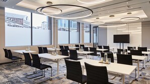 A conference room with tables, chairs, a large screen, and a view of buildings through large windows. - Cardiff Marriott Hotel (Cardiff)