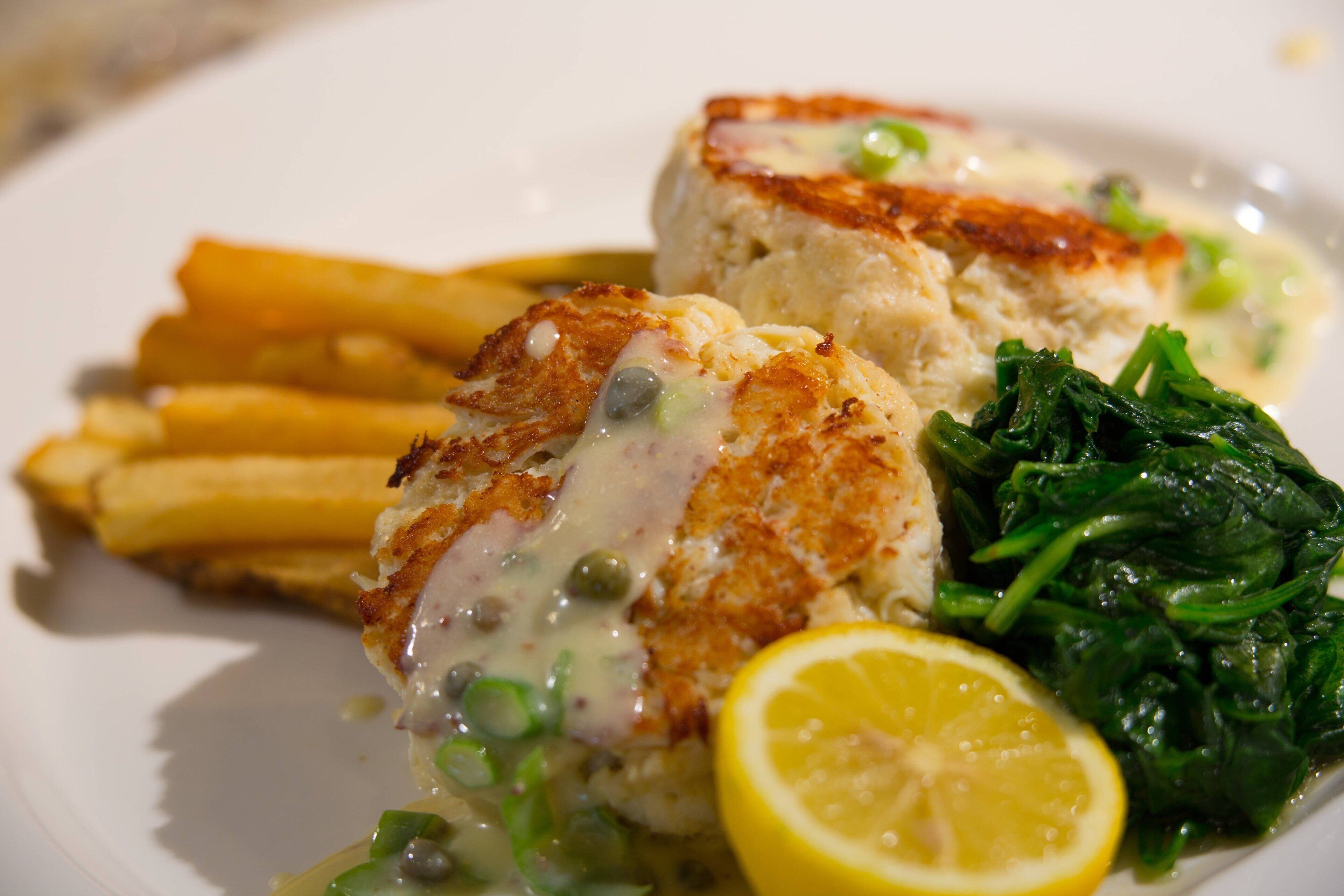 Un plato de comida con un trozo de pescado, papas fritas, espinacas y una cuña de limón.