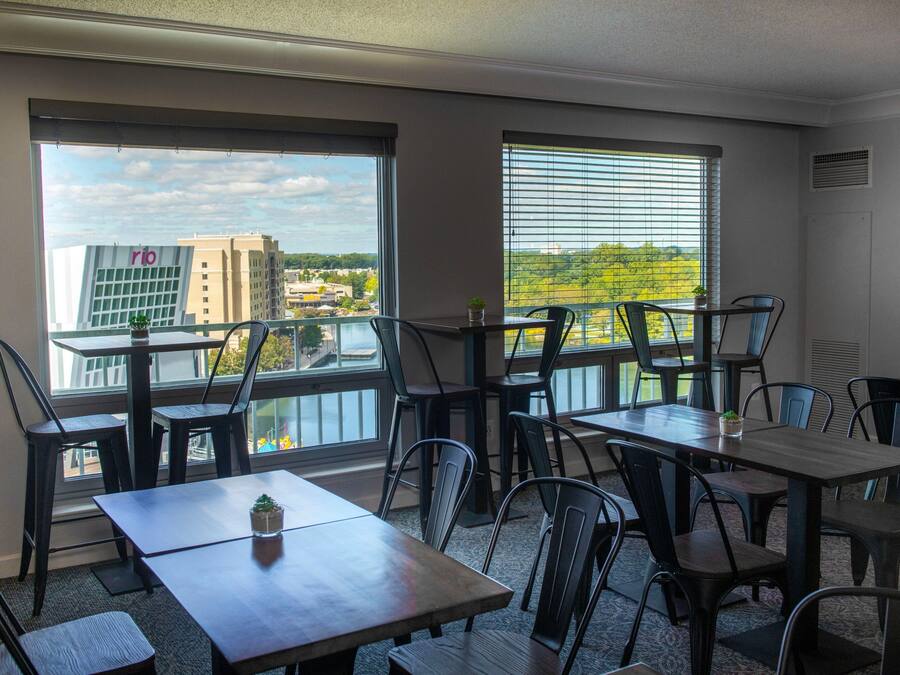 A dining area with tables and chairs, large windows with blinds, and a view of buildings and trees.