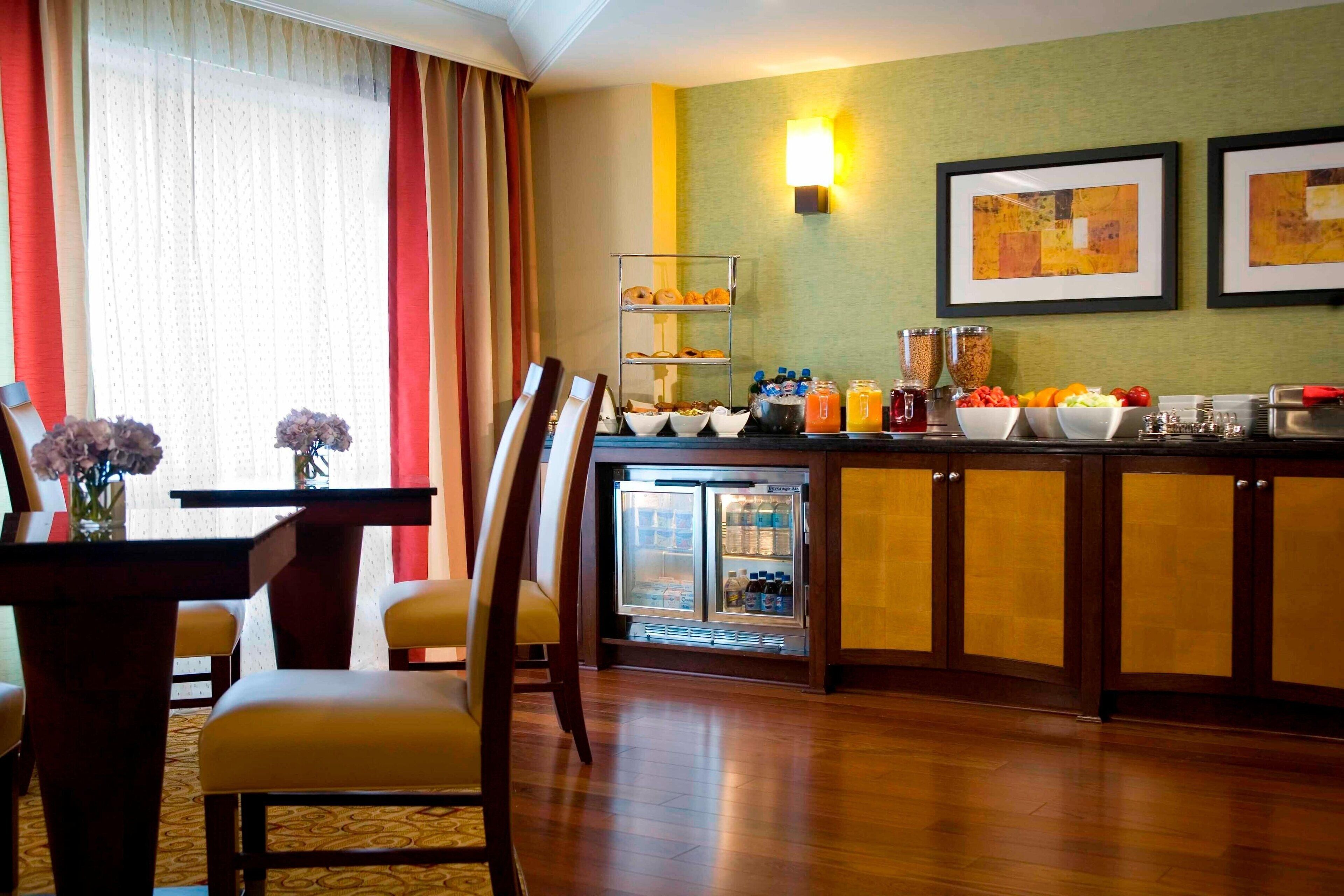 Une chambre d’hôtel dotée d’un coin repas comprenant une table et des chaises, d’un réfrigérateur contenant des boissons et d’œuvres d’art accrochées au mur.