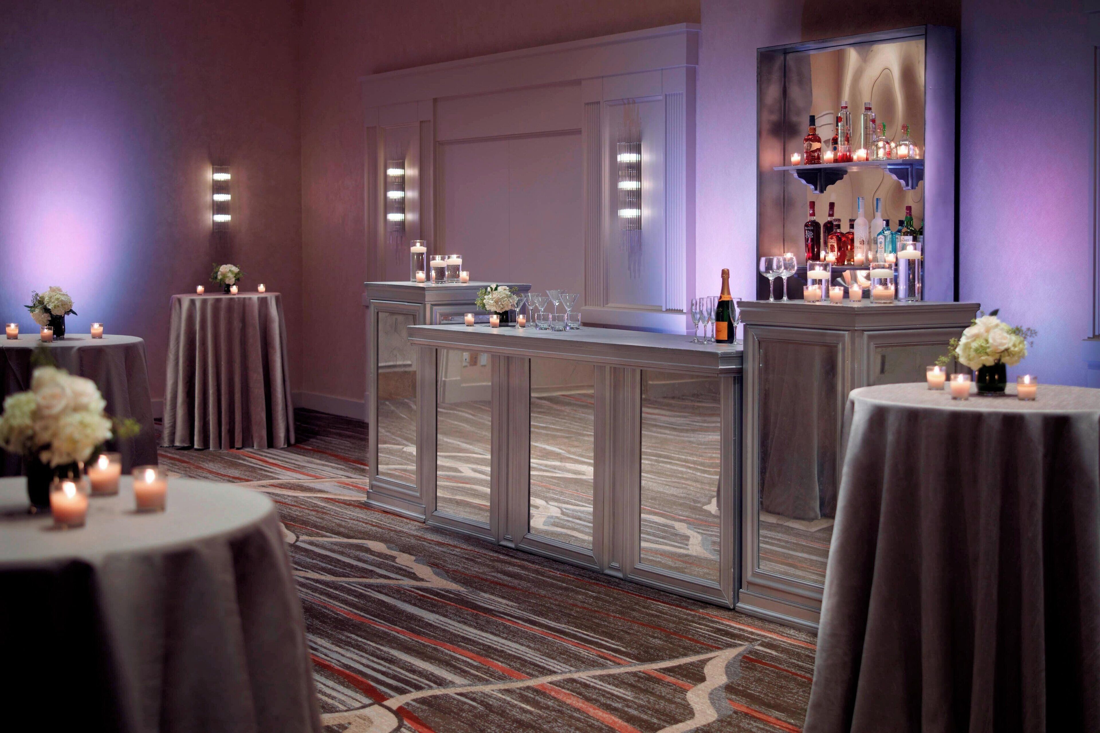 Vestíbulo de hotel con mesas redondas, velas y una zona de bar con botellas y copas.