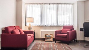 Une chambre d’hôtel moderne avec deux fauteuils rouges, une petite table d’appoint en bois sur laquelle se trouve une lampe, une grande fenêtre avec des rideaux légers et un tapis à rayures sur le sol.
