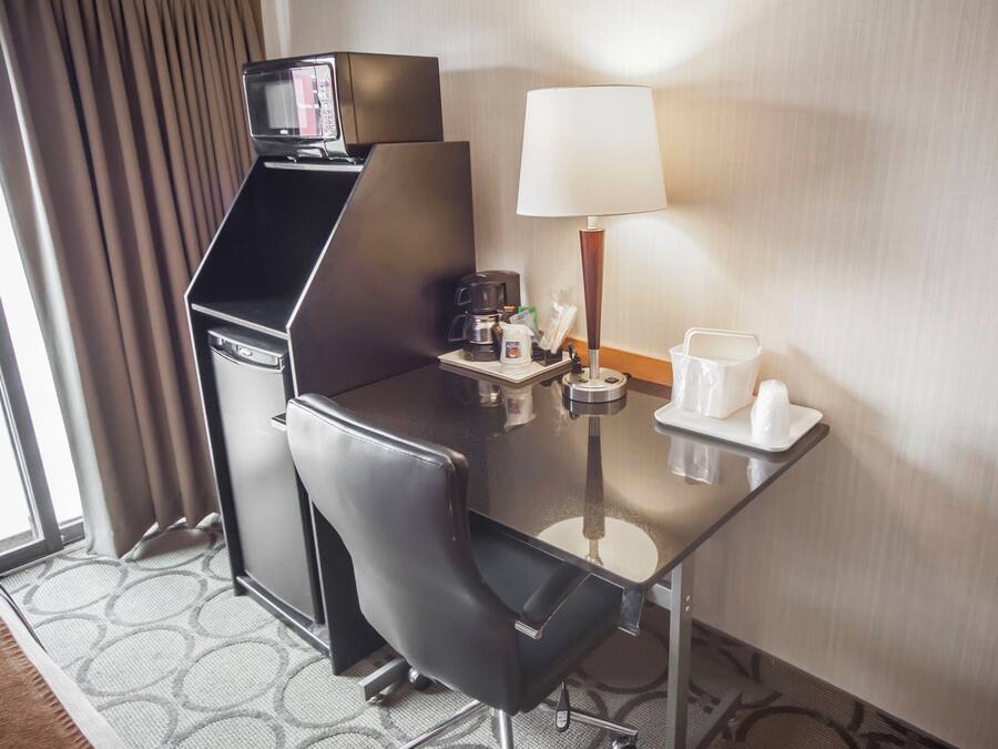 A hotel room with a compact desk, a coffee maker, a lamp, and a small table with cups and a bottle.