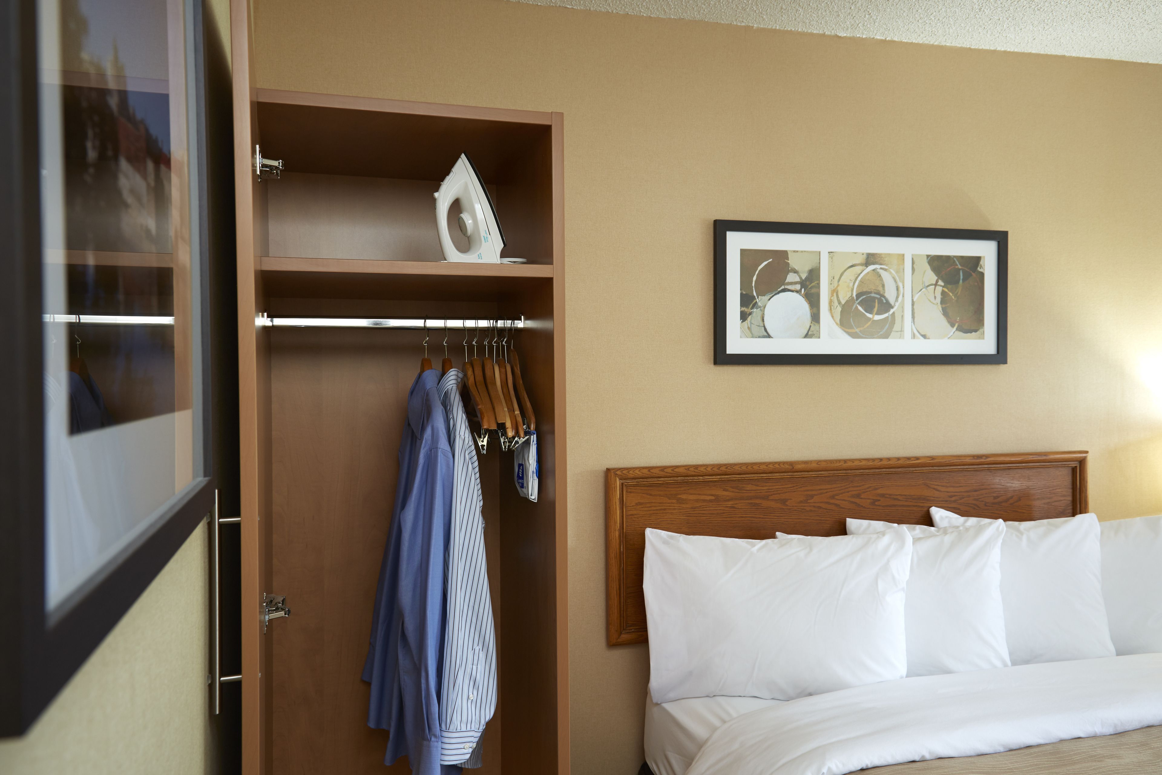 A hotel room with a wooden wardrobe, a bed with white bedding, and a framed abstract art piece on the wall.