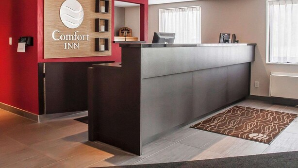 Un espace d’accueil d’hôtel avec un mur rouge, un comptoir de réception en bois et un présentoir gris.