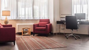 Une chambre d’hôtel avec un fauteuil rouge, une chaise de bureau noire, un bureau avec un ordinateur et une lampe.