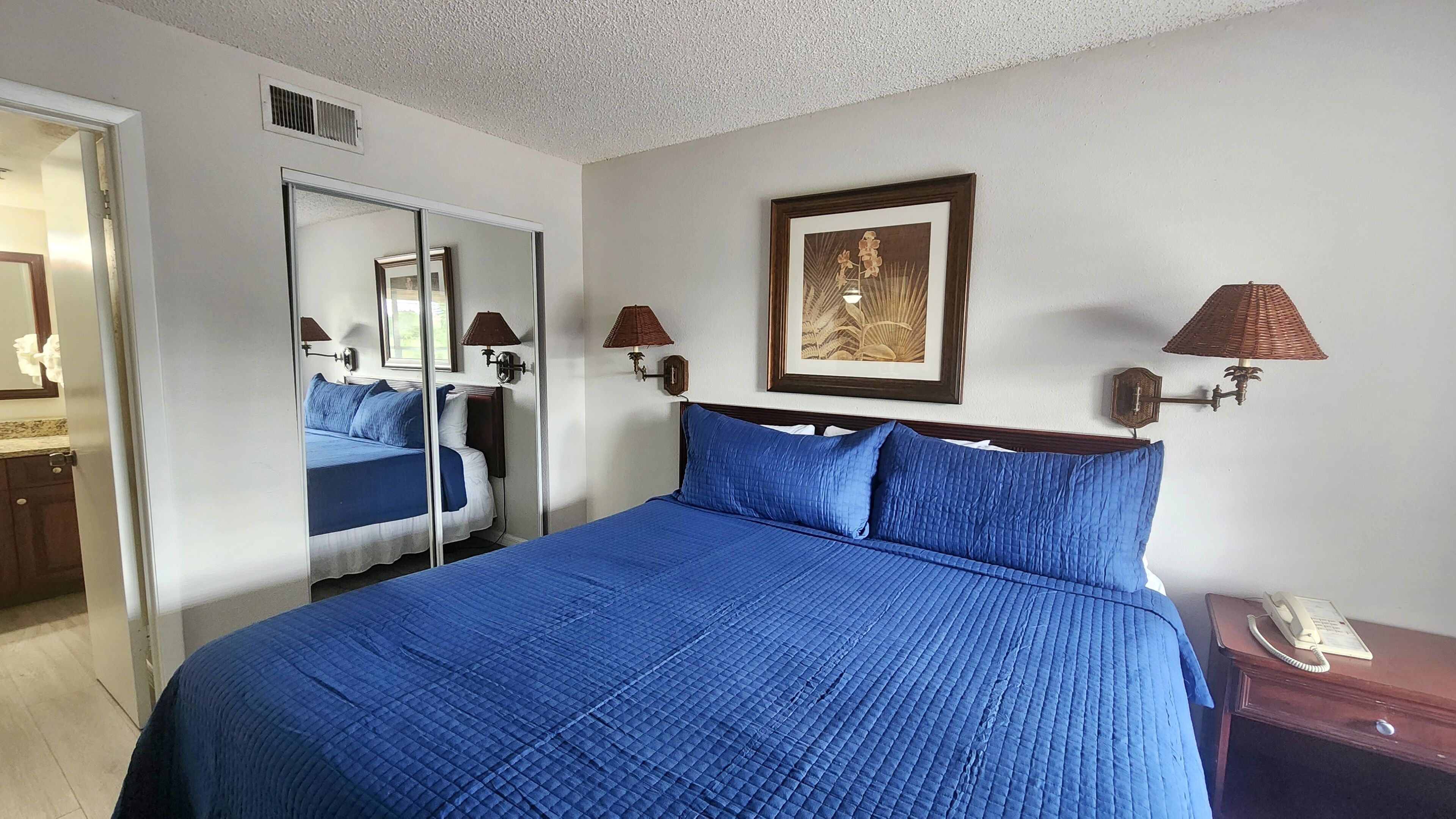 A hotel room with a large bed, a mirror, two wall-mounted lamps, a framed picture, and a bedside telephone.