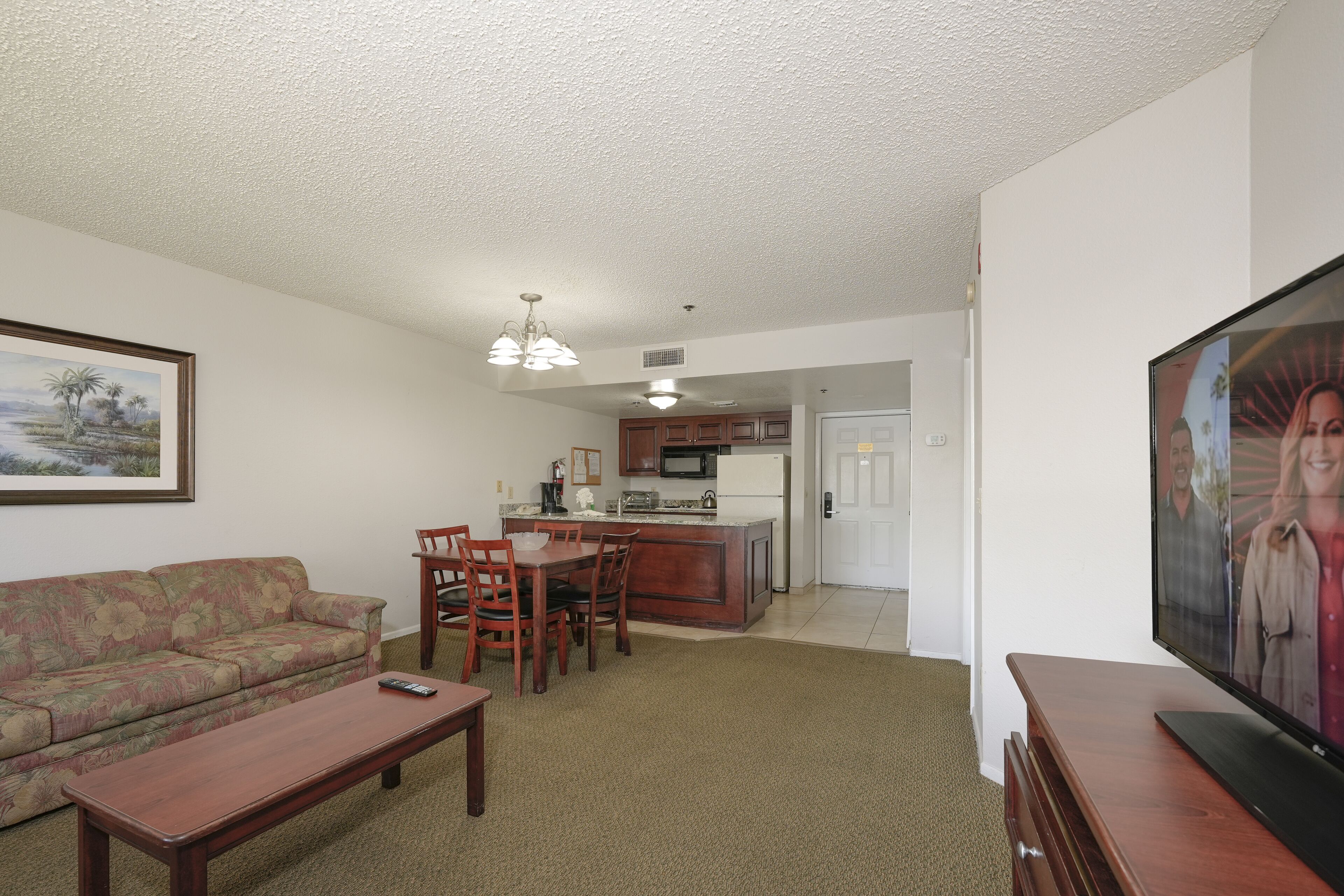 A living room with a sofa, a coffee table, and a television. There is a kitchen area with a dining table and chairs.