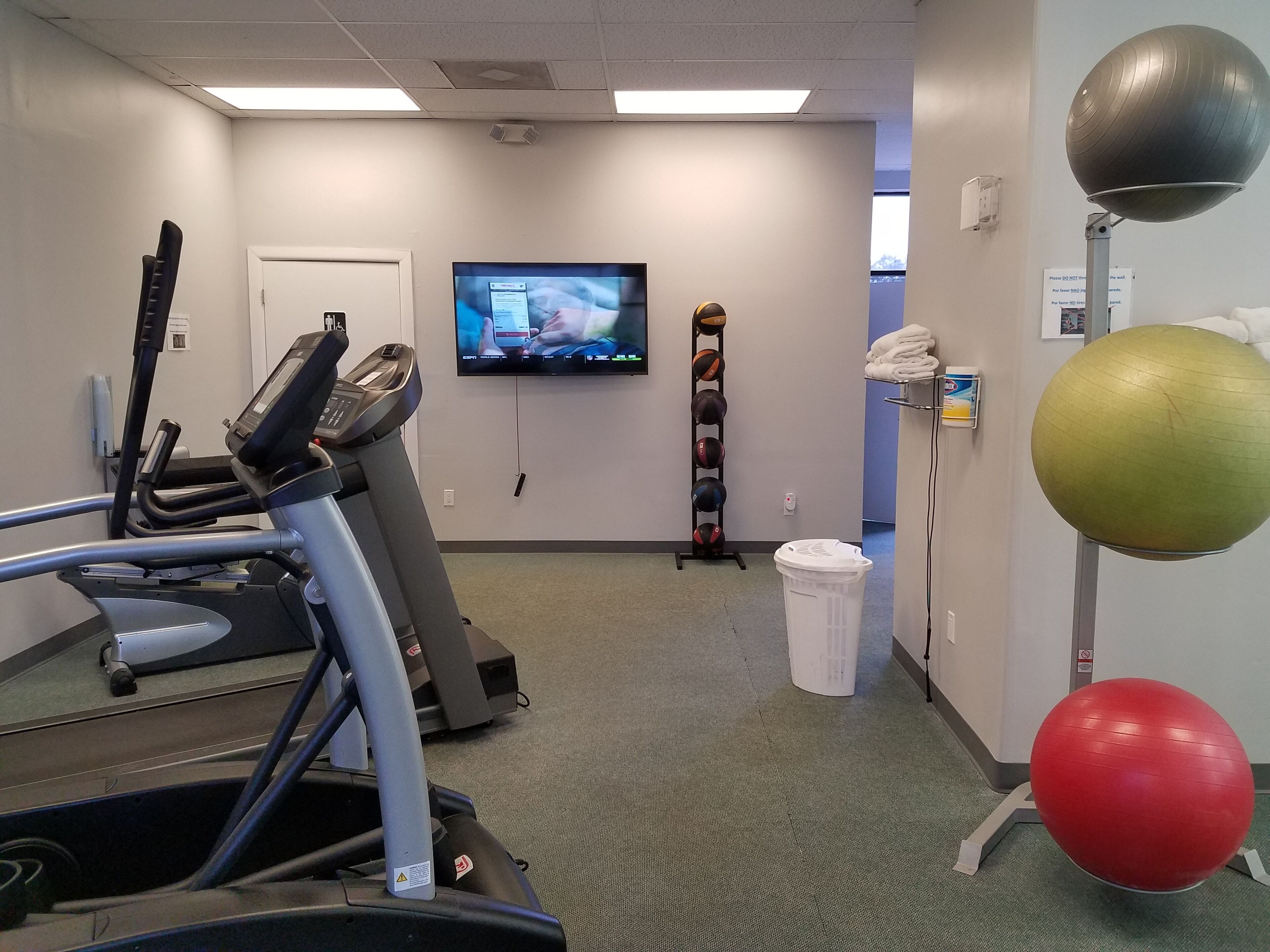 Una sala de fitness amb cintes de córrer, televisió, pilotes d'exercici i una cistella de roba.