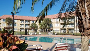 A swimming pool with lounge chairs and umbrellas, surrounded by palm trees and a multi-story building with balconies. - Parc Corniche Condominium Suite Hotel (Orlando)