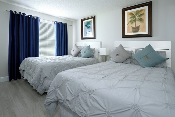 A hotel room with two beds, a large window with blue curtains, and a framed picture of a palm tree on the wall.
