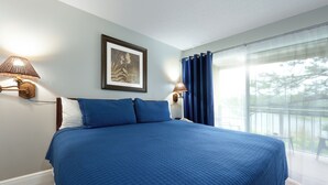 A bedroom with a large bed, two wall-mounted lamps, a framed picture, and a view of the outdoors through a window with blue curtains. - Parc Corniche Condominium Suite Hotel (Orlando)