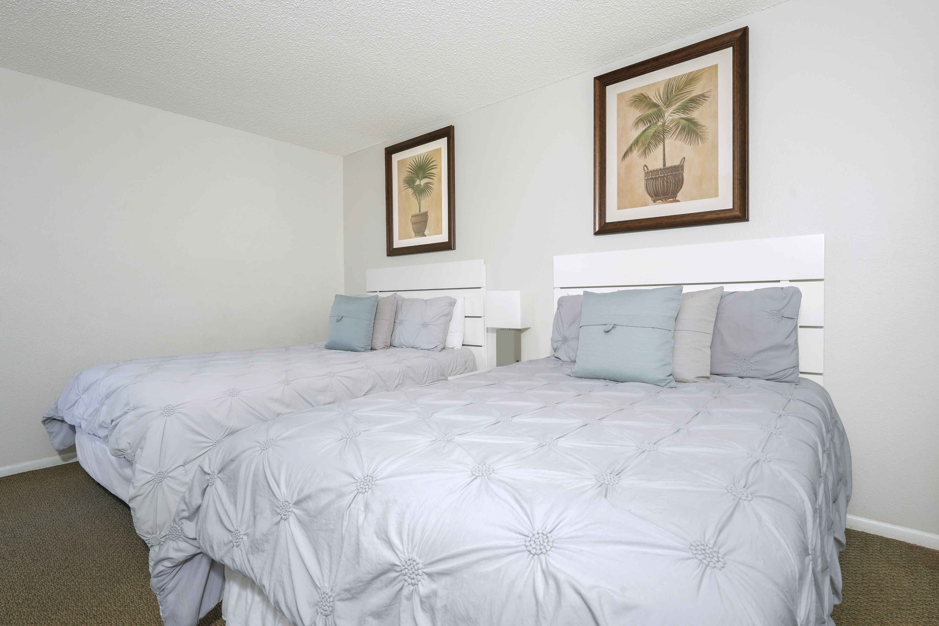 Un lit double avec une tête de lit blanche et une couvre-lit à motifs floraux gris. Deux tableaux encadrés sont accrochés au mur au-dessus du lit.