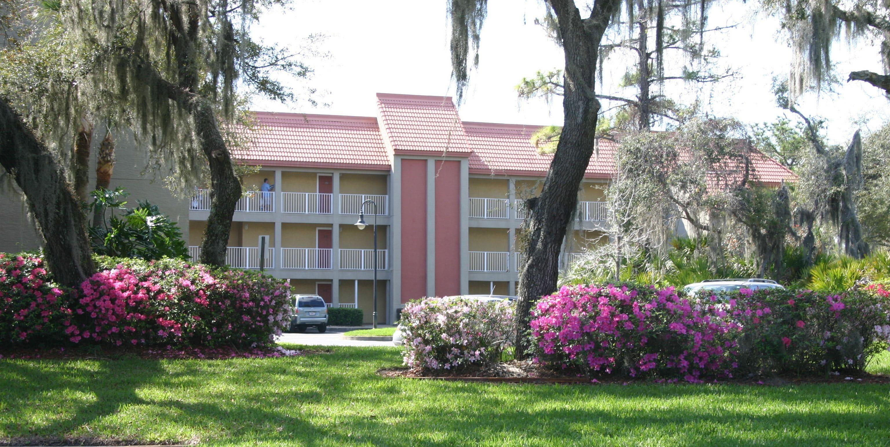 Un motel à plusieurs étages, avec un toit rouge, des balcons et entouré d’une végétation luxuriante et de fleurs roses.