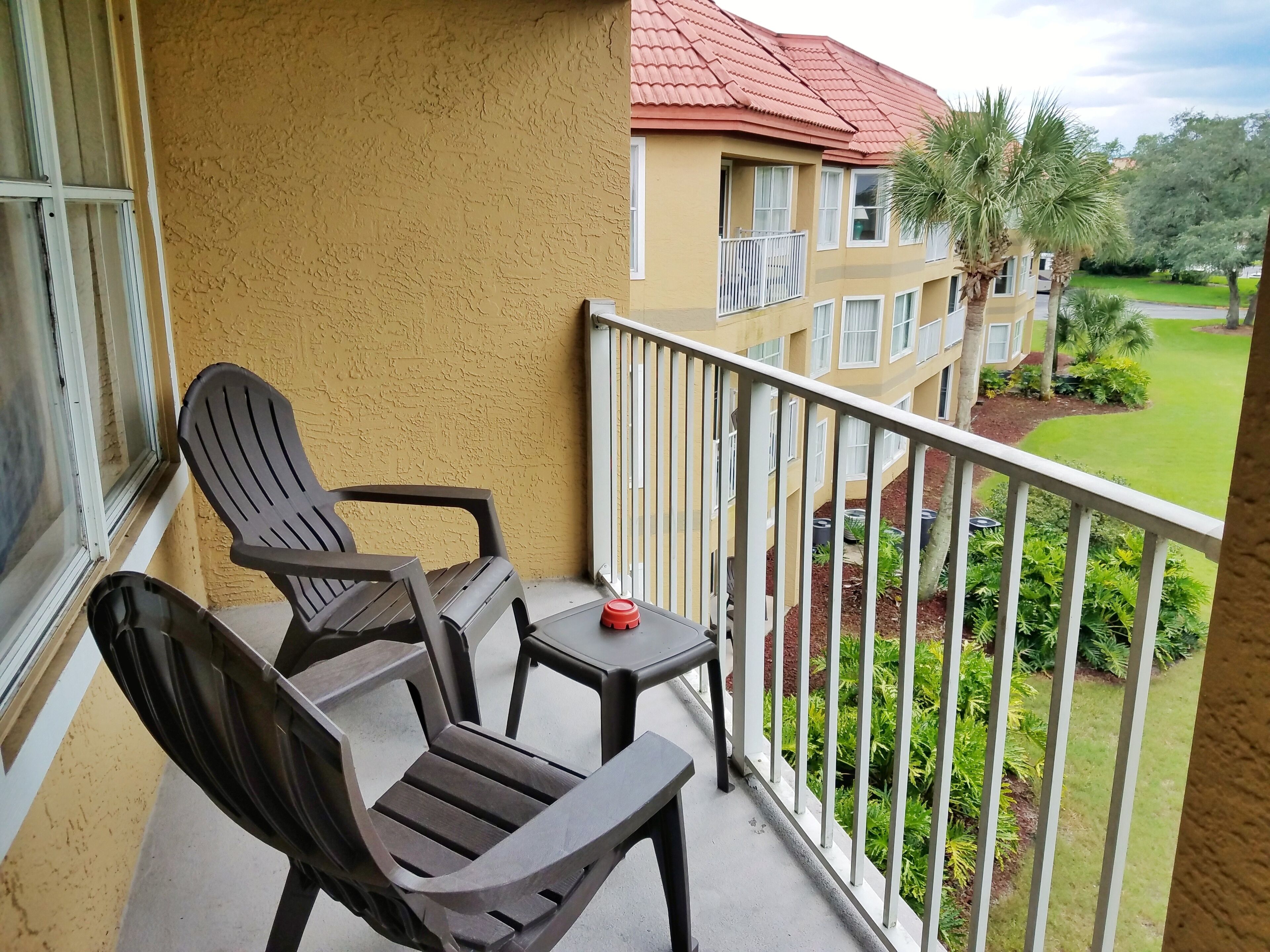A balcony with two chairs and a small table, overlooking a well-maintained lawn and a building with a red roof.