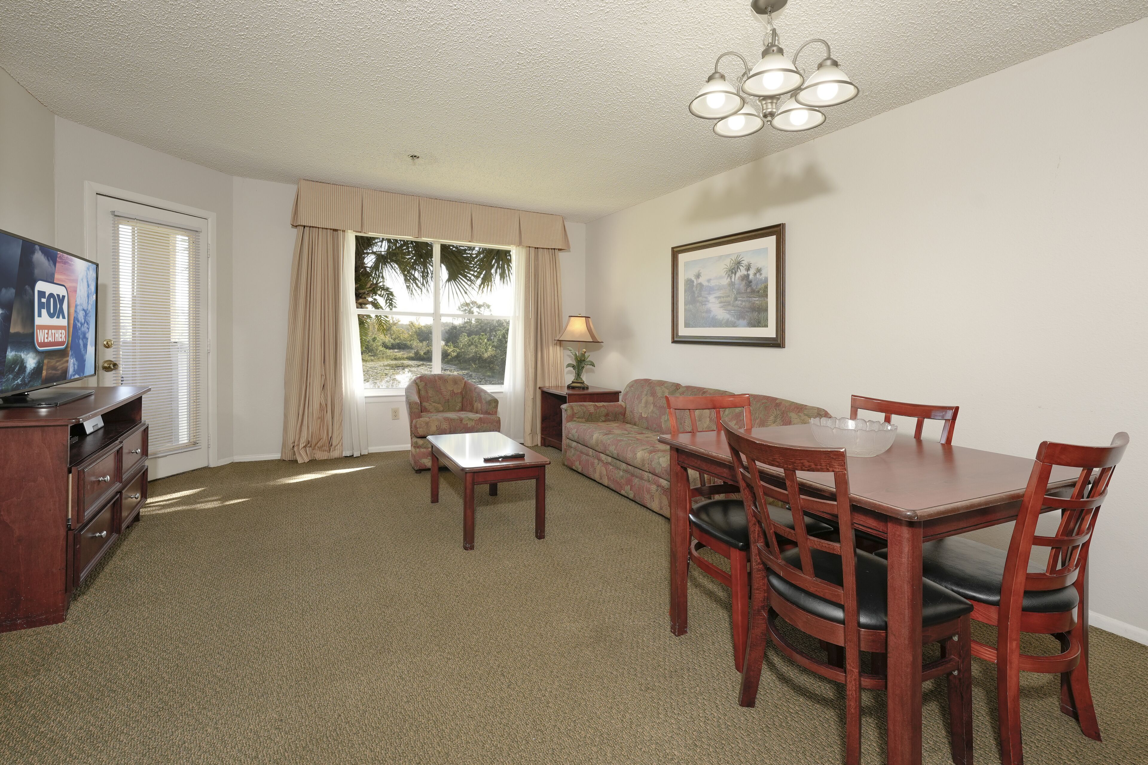 A furnished living room with a television, a sofa, a dining table with chairs, a lamp, and a window with curtains.