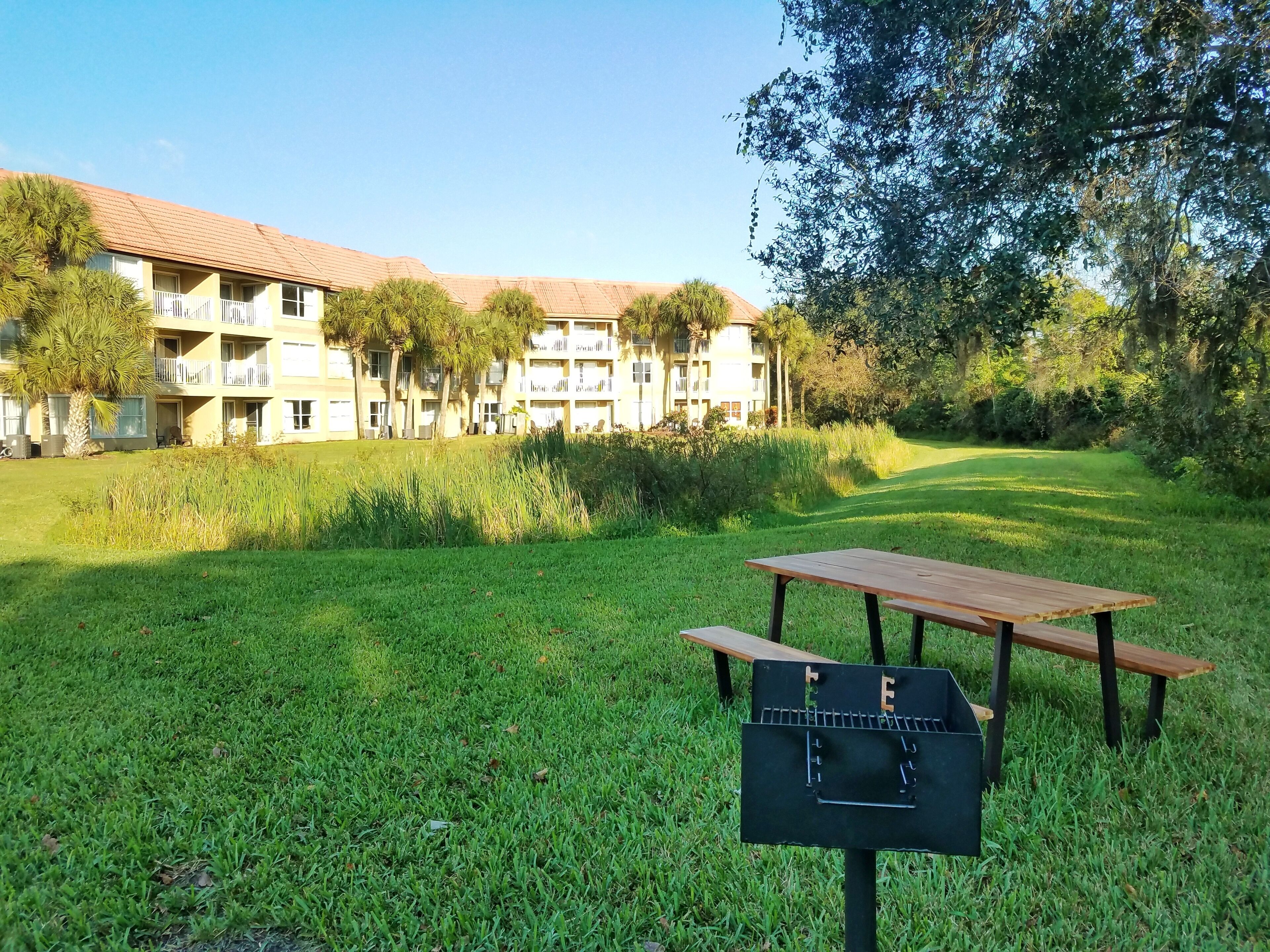 Un espacio al aire libre con césped, una mesa de picnic y una parrilla, rodeado de abundante vegetación y un edificio con varios balcones.