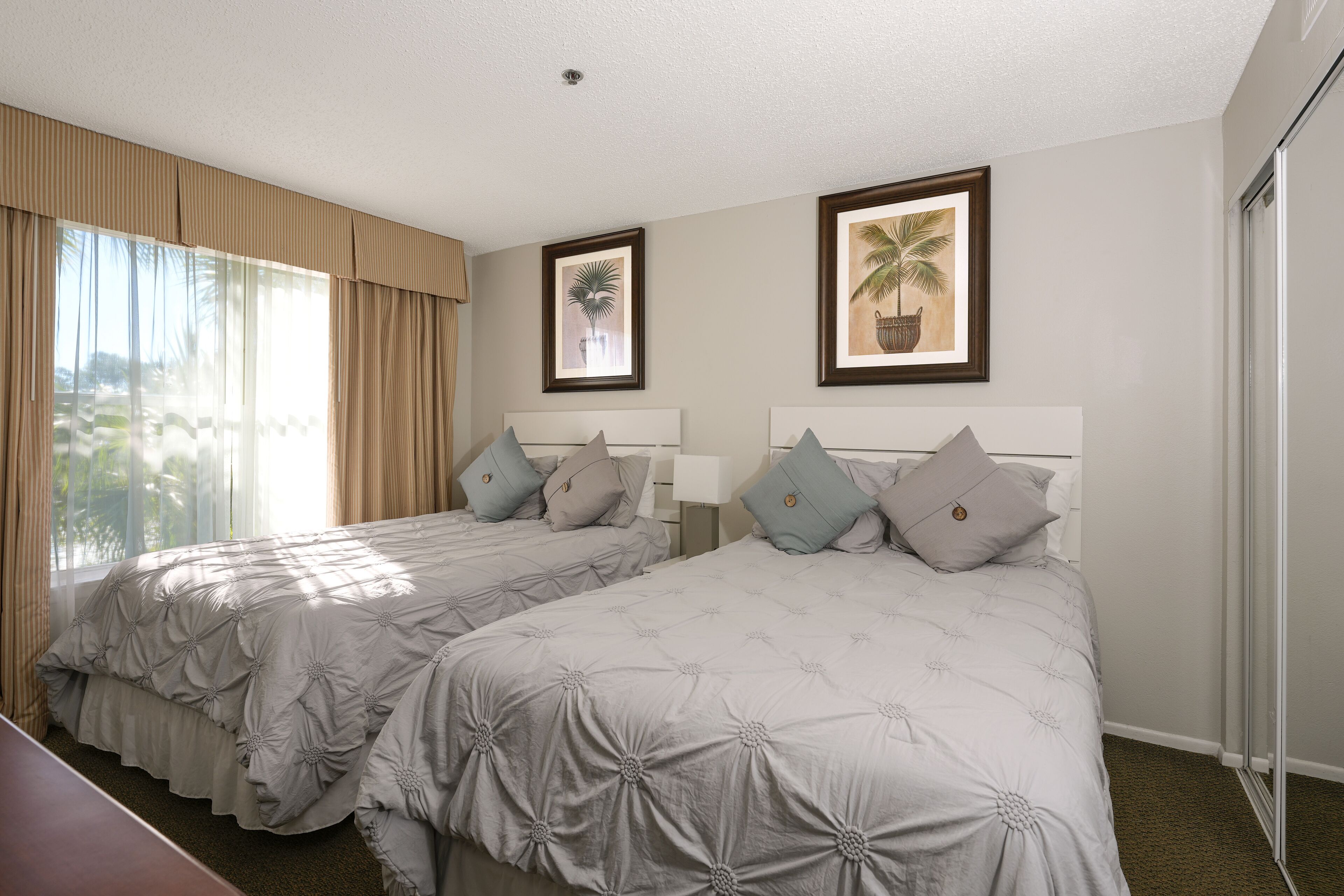 A hotel room with two beds, a large window with curtains, and two framed pictures on the wall.