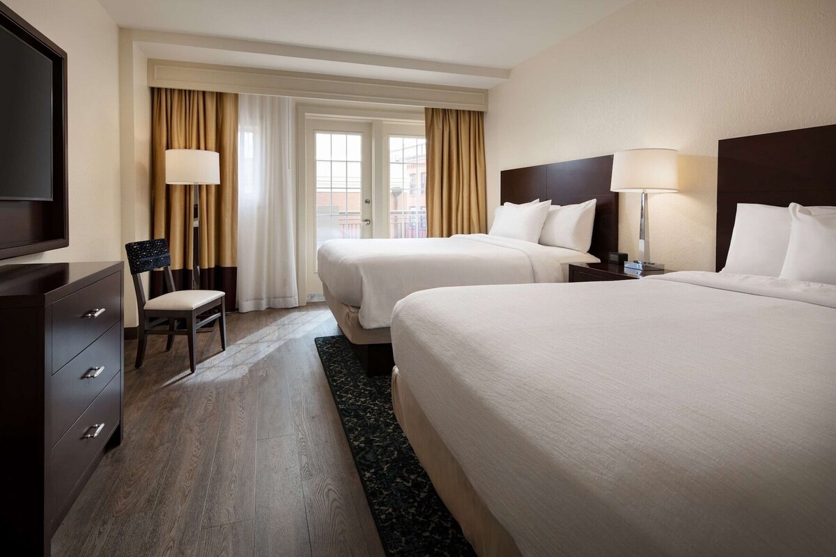 Ein Hotelzimmer mit zwei Betten, einem Schreibtisch, einem Stuhl, einem Fernseher und einem Fenster mit Vorhängen.