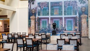 A dining area with tables and chairs, a mural of a building, and a fountain. - Embassy Suites by Hilton New Orleans (New Orleans)