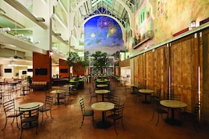 A spacious indoor atrium with multiple dining tables and chairs, a high ceiling with a mural, and a wooden wall on one side.