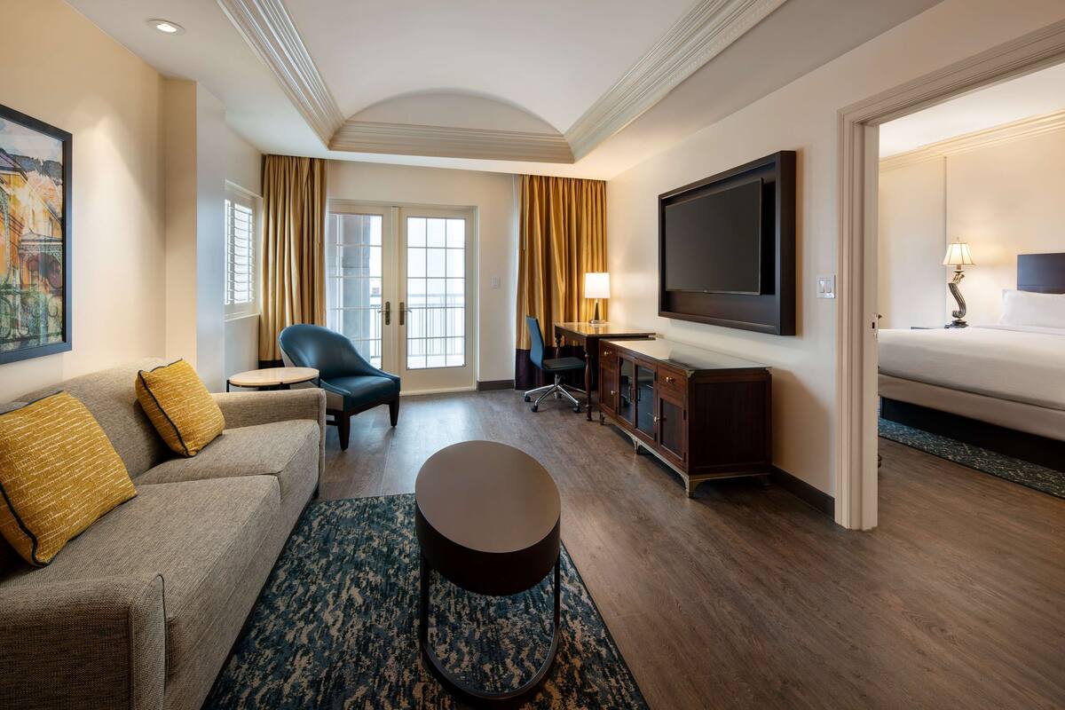 Een hotelkamer met een bank, een ronde salontafel, een aan de muur bevestigde televisie, een bed en een bureau met een lamp.