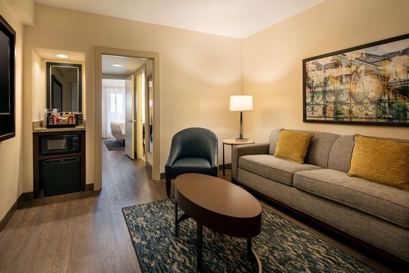 A hotel room with a kitchenette, a sofa, a chair, a small table, a lamp, and a painting on the wall.