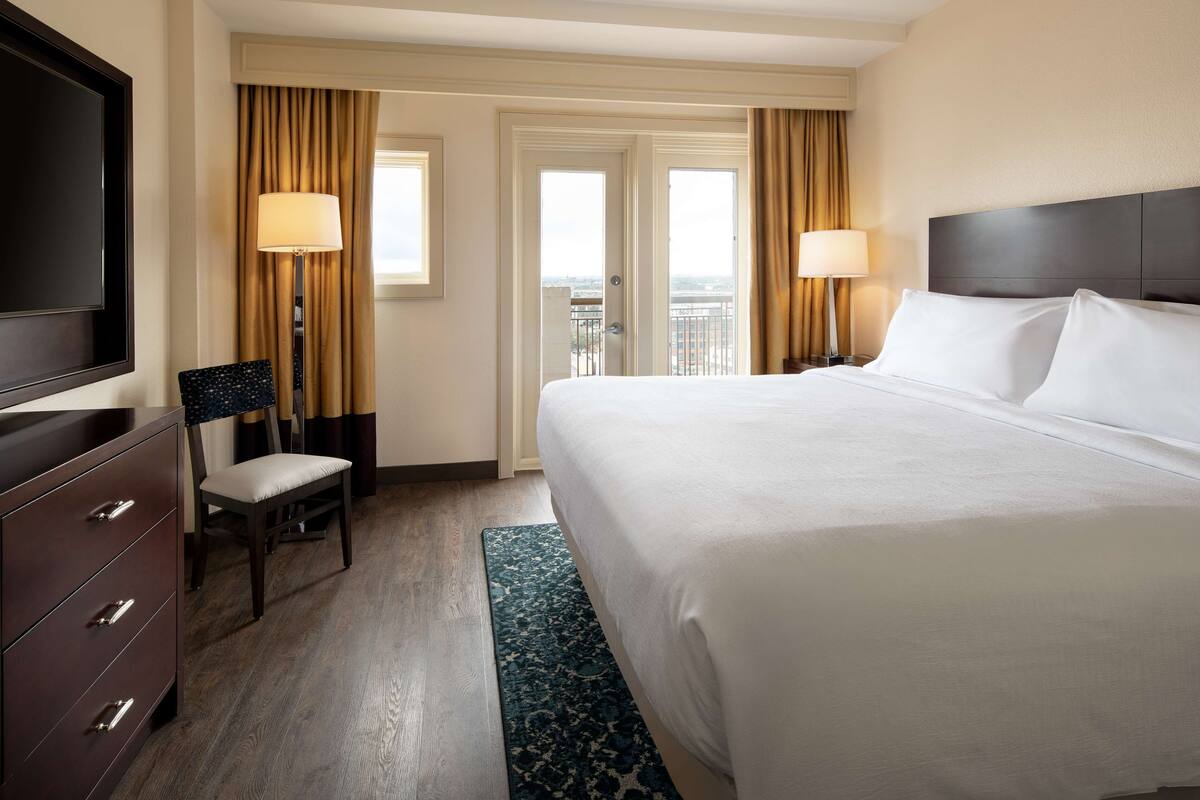 Een hotelkamer met een groot bed, een televisie, een bureau met lades, een stoel en uitzicht op de stad.