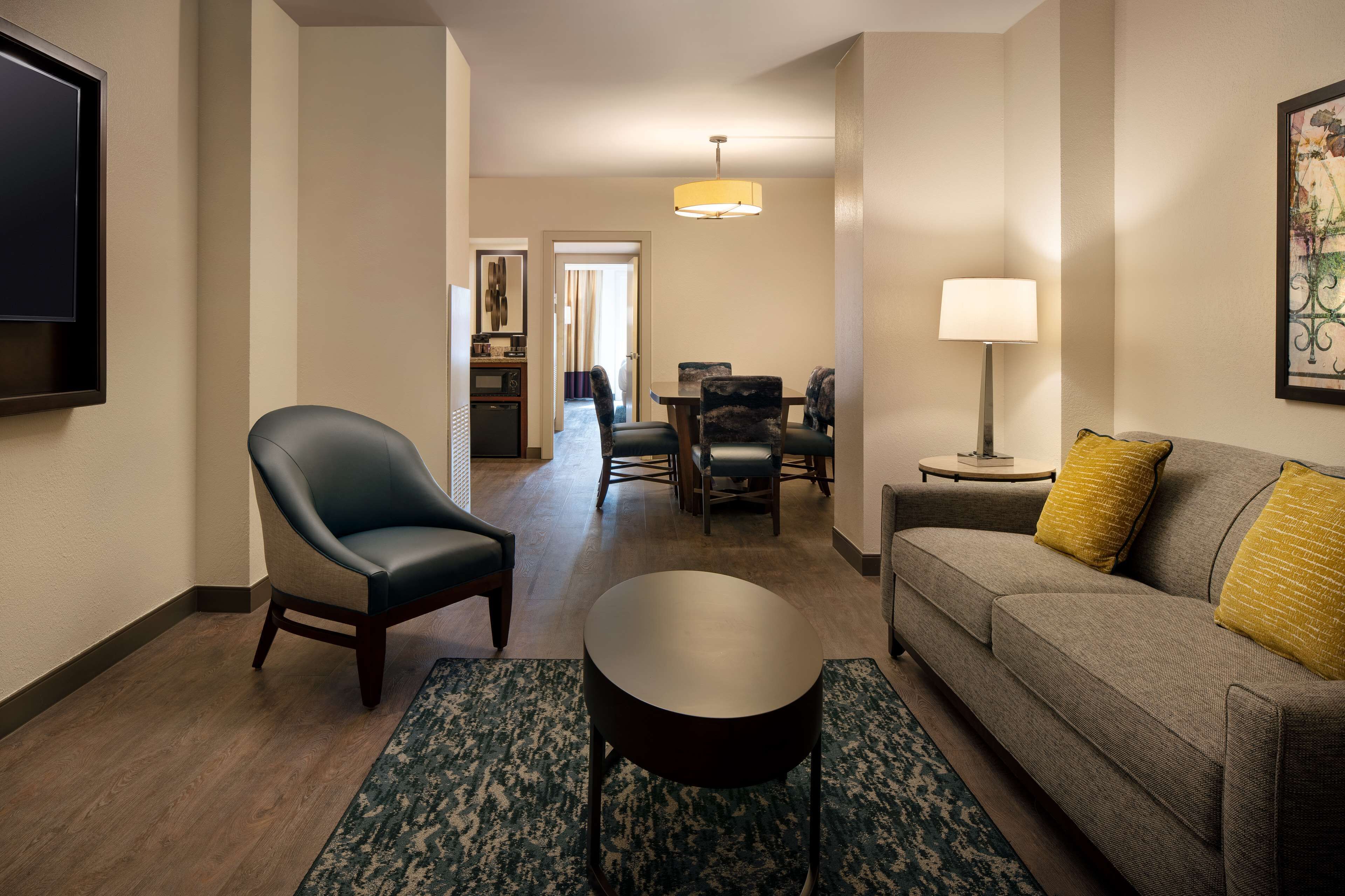 Una habitación de hotel con sofá, sillón, mesa de comedor y televisión montada en la pared.