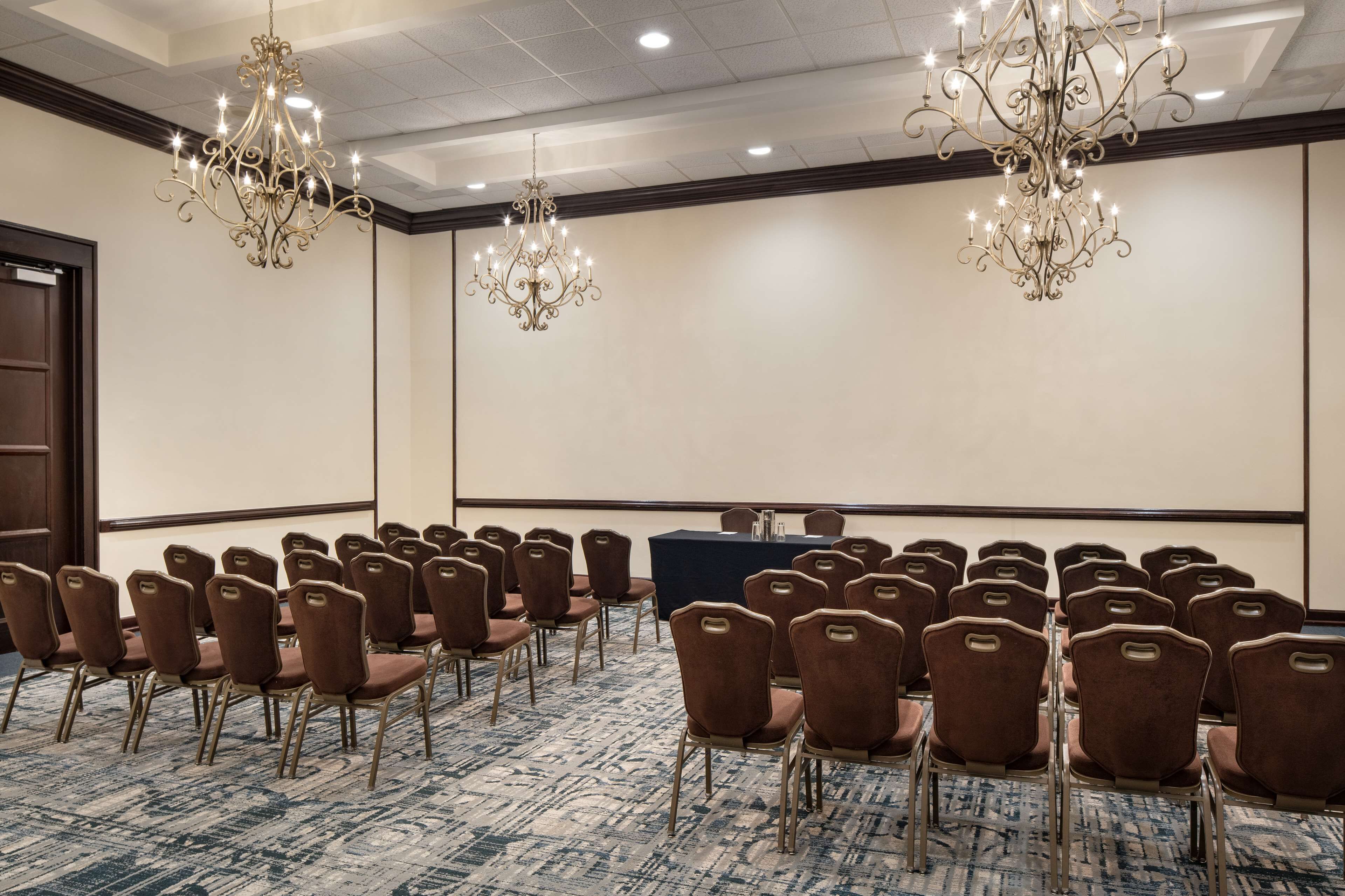 Un salón de conferencias con filas de sillas marrones, una mesa larga y dos candelabros.
